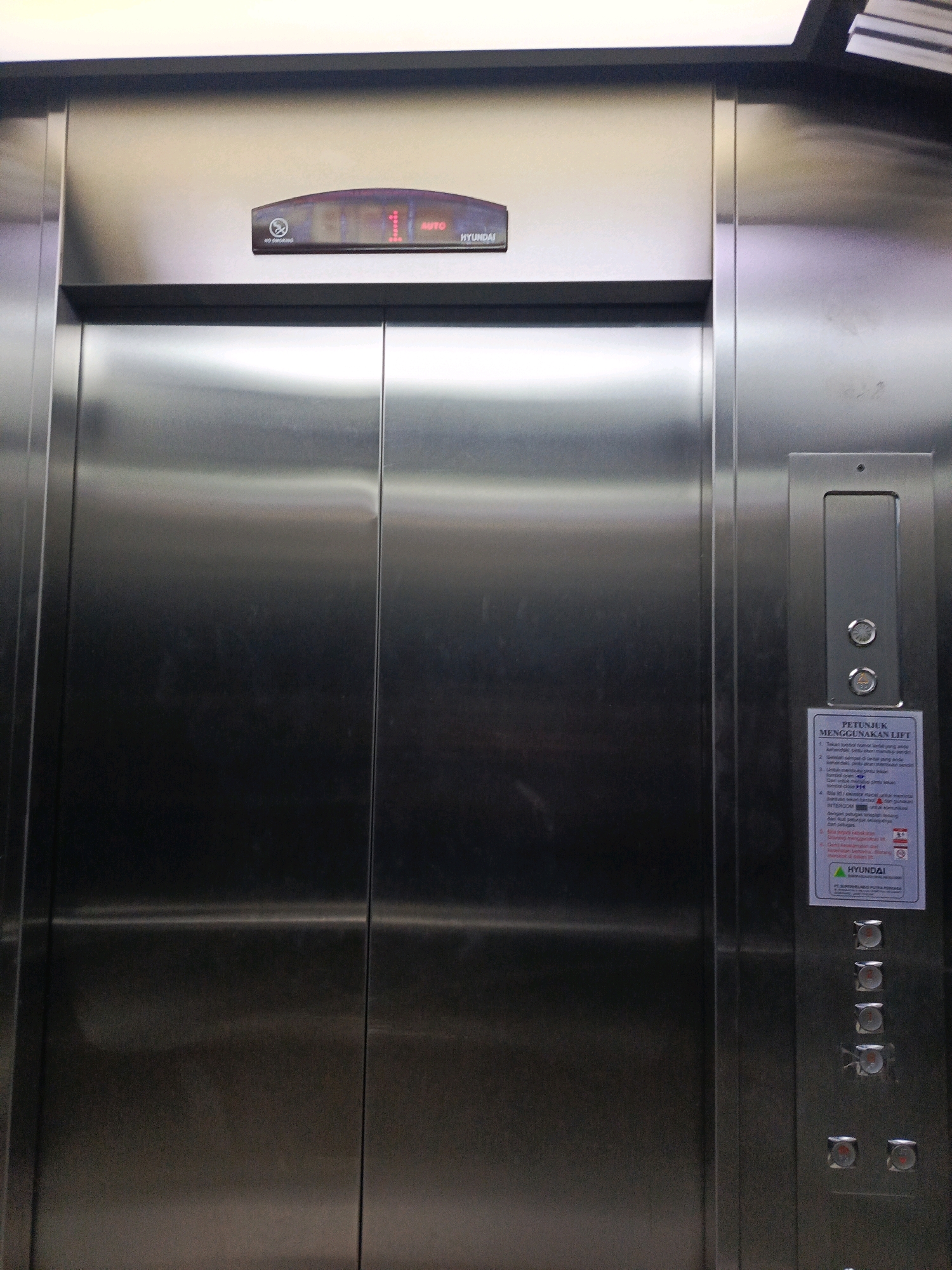 The image shows the interior of a stainless steel elevator with two doors. Above the doors, a digital display shows "1" and "AUTO", indicating the current floor and operating mode. To the right of the doors, a panel with buttons and a user manual is visible. The user manual is in Indonesian and provides instructions on how to use the lift. The elevator appears to be in a public building, likely in Tegal, Indonesia, given the language on the manual.