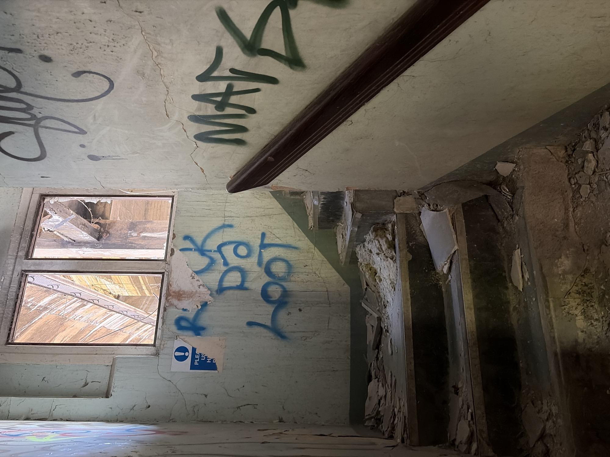 The image shows the interior of a derelict building, likely located in Mid Suffolk, United Kingdom, although this cannot be forensically confirmed. The room has two broken windows, graffiti on the walls including the word "MATA", and a damaged doorway. The floor is littered with debris. There are no people visible.