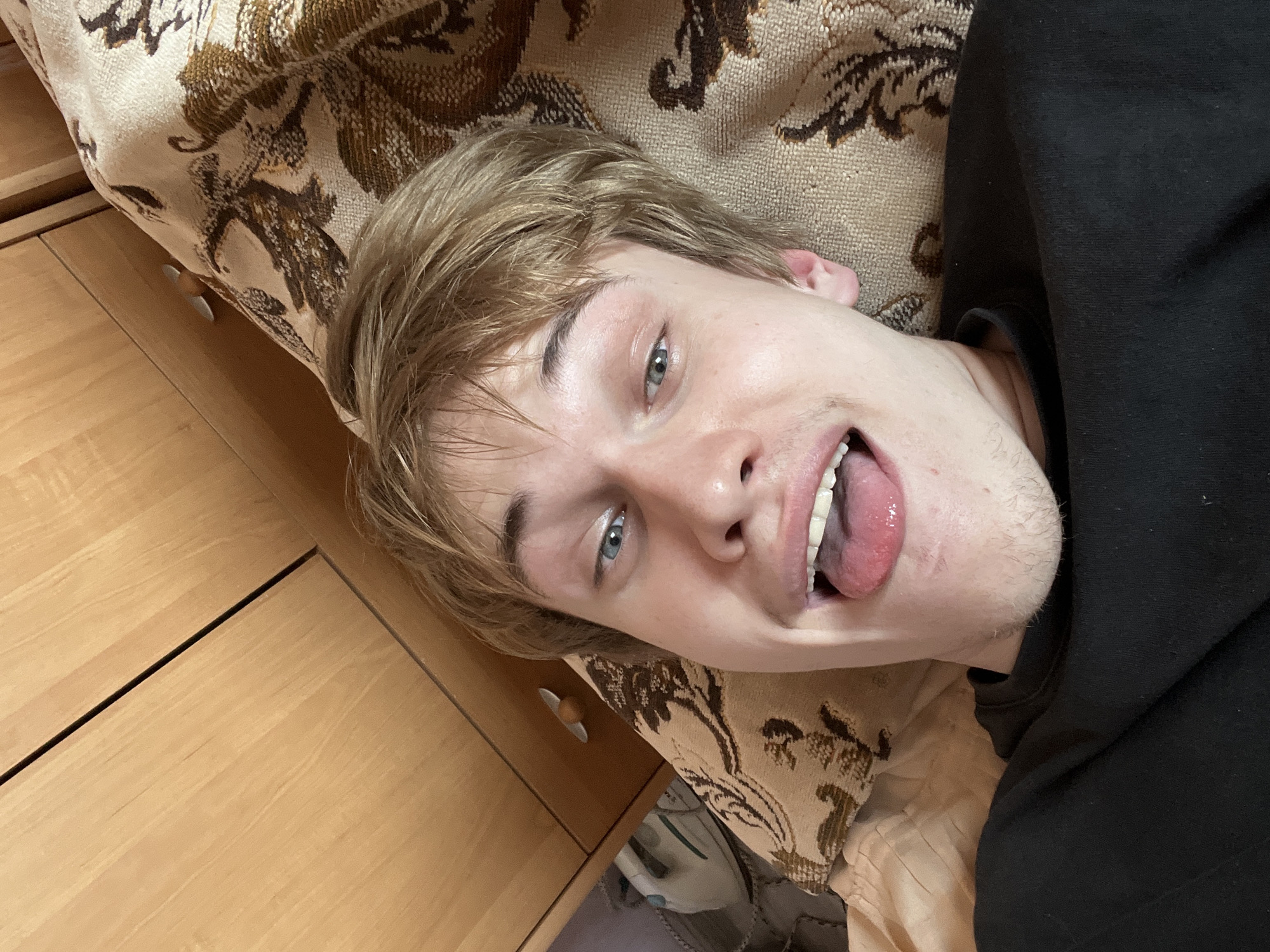 The image shows a close-up, overhead view of a young Caucasian male lying on a patterned textile, likely a bedspread. He has light brown hair and blue eyes, and he is sticking his tongue out with his mouth open. He is wearing a black long-sleeved shirt. In the background, there is a wooden cabinet with drawers, and an object that appears to be an iron is visible on the left side of the frame. The overall environment appears to be indoors, in a domestic setting. There is no specific indication of the image being taken in the city of Gari, Russia.