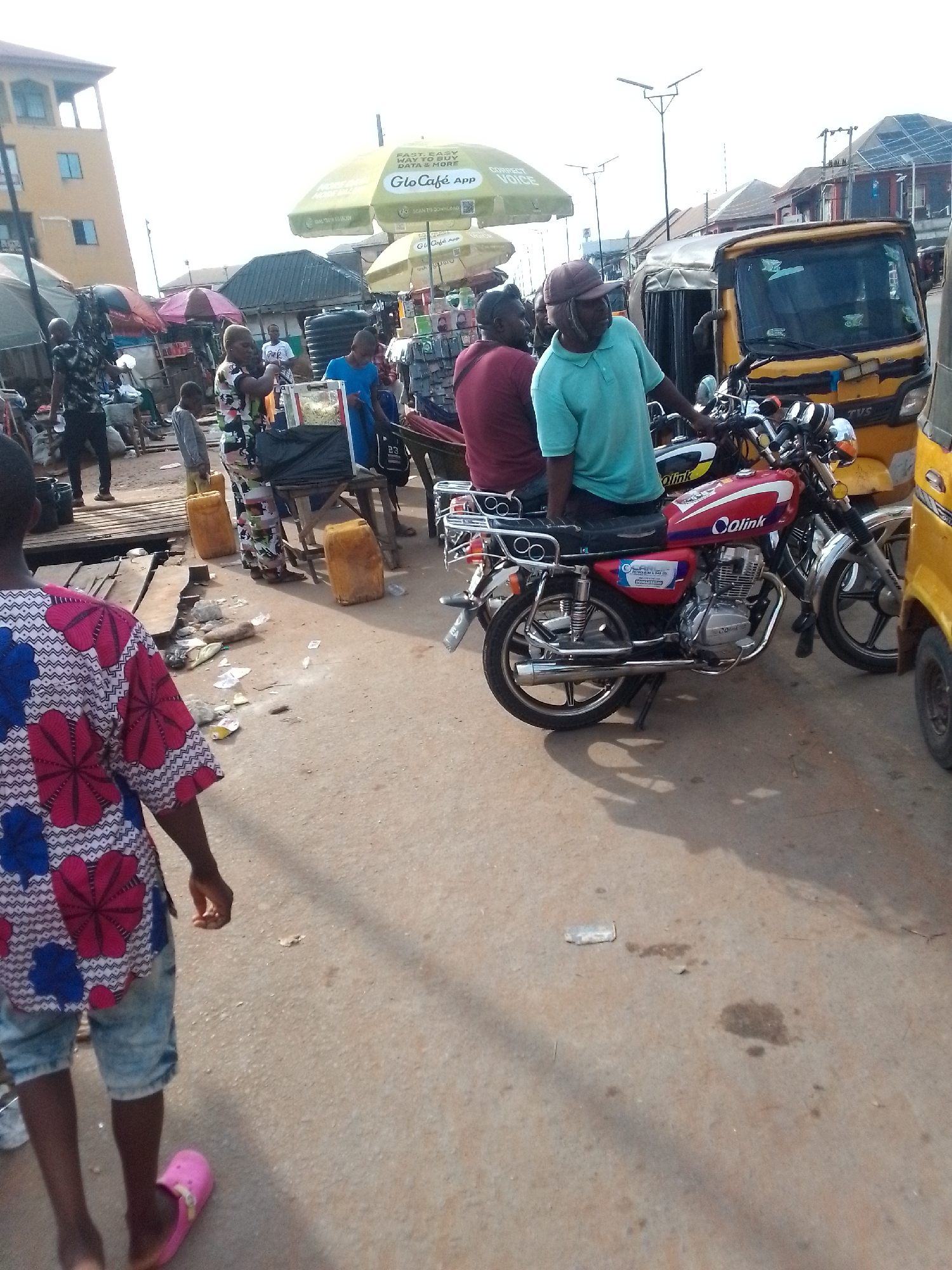 The image depicts a street scene in what appears to be a market area of Orlu, Nigeria. The ground is paved and contains litter. Several people are present, including a child walking away from the camera in the foreground wearing a patterned shirt and pink sandals. In the background, people are gathered around vendor stalls, indicated by yellow jerrycans and umbrellas branded with “Glo Café App.” A red and silver motorcycle labeled "Qlink" is parked near a yellow three-wheeled vehicle, known locally as a "Keke Napep." Two men are sitting on the motorcycle.