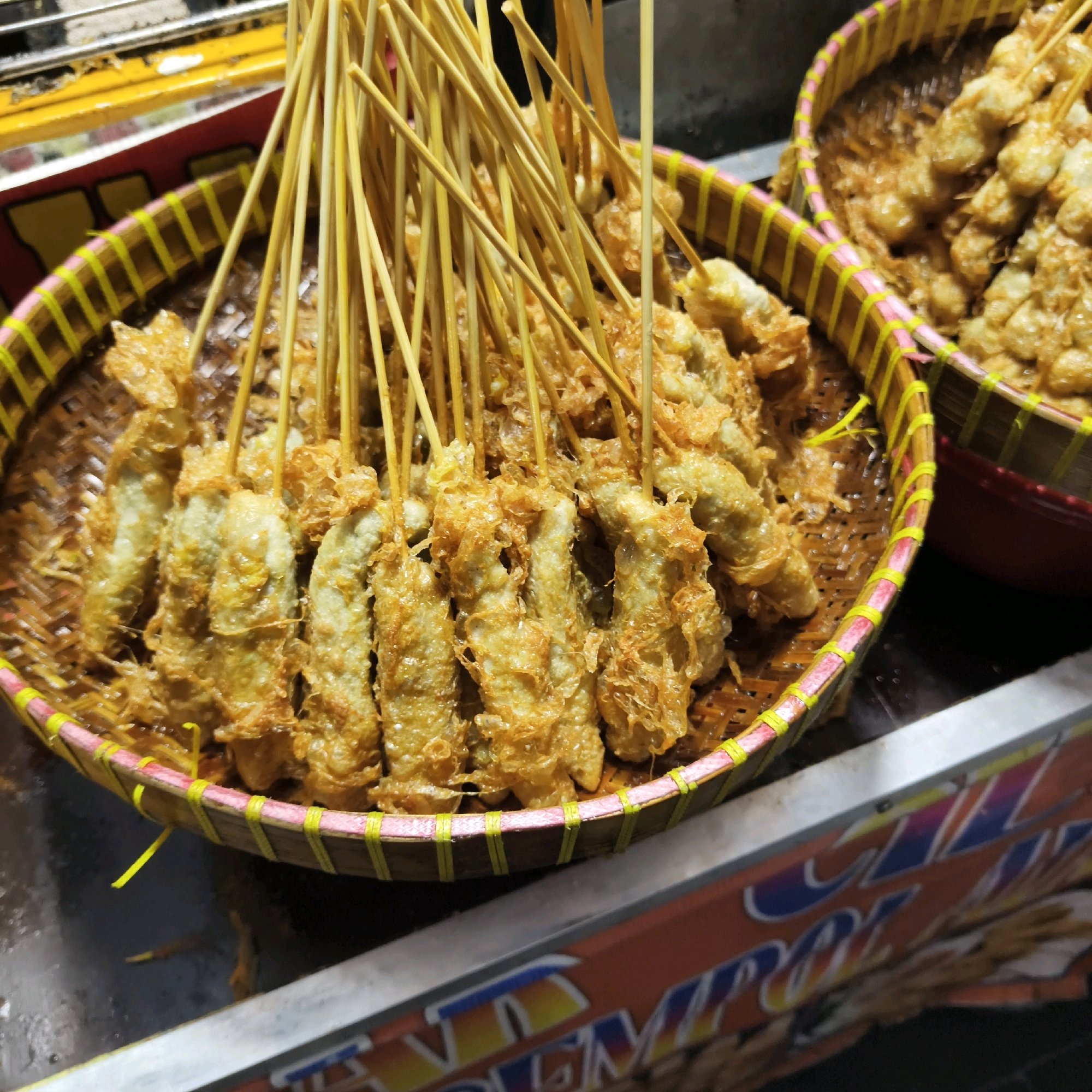 The image shows a close-up of a woven basket filled with deep-fried spring rolls on skewers, a common street food in Indonesia. The spring rolls are golden brown and appear crispy, with some strands of fried batter visible. The basket has a decorative trim of pink and yellow woven material. In the background, another basket of spring rolls is partially visible, as is a red bowl. A brightly colored sign with text in Indonesian is also visible in the foreground. The scene suggests a food stall or market in West Jakarta, Indonesia, likely a busy, casual food vendor. The lighting is warm and somewhat artificial, typical of street food settings.
