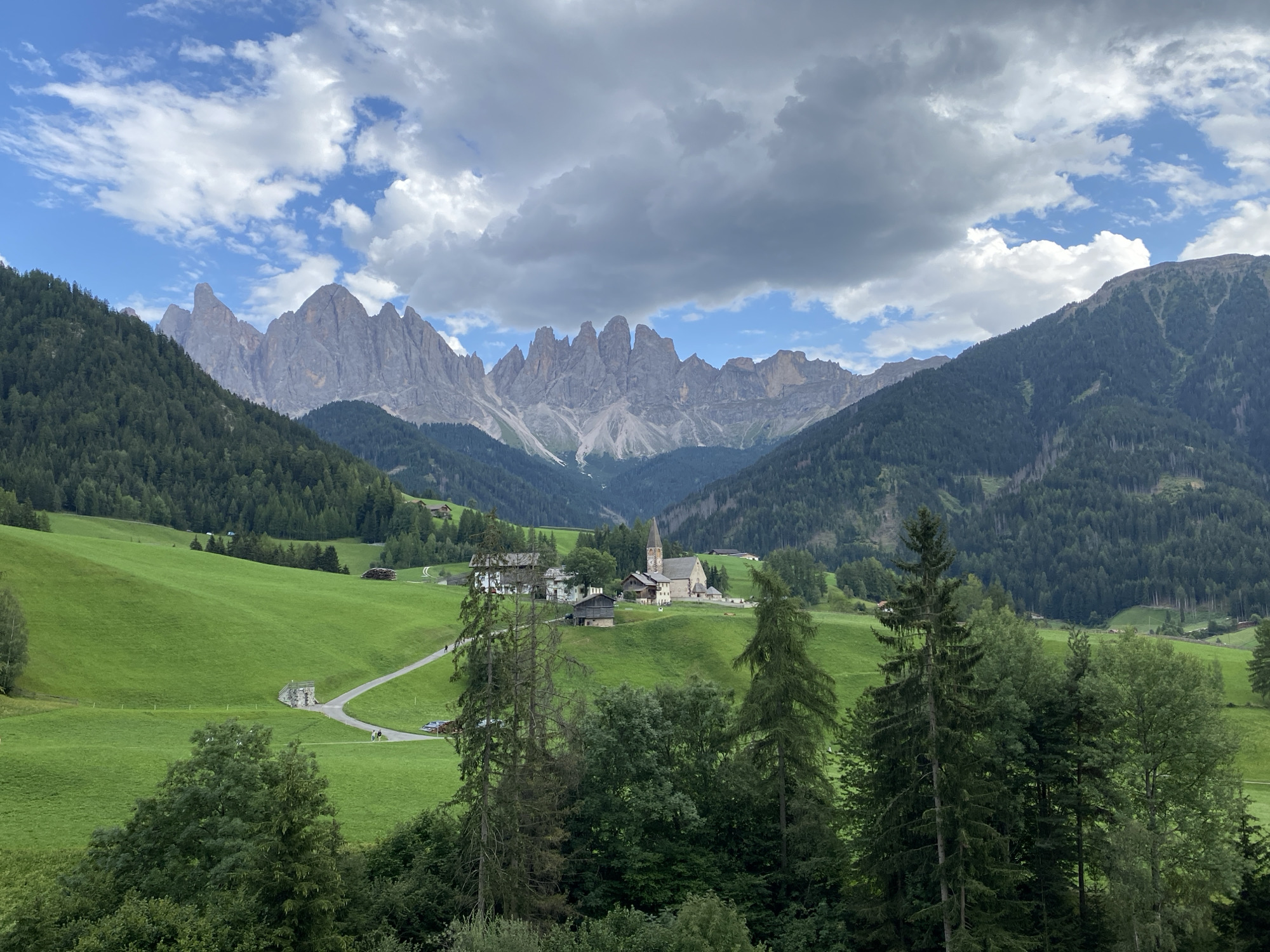 The image depicts a scenic view of St. Magdalena (Santa Maddalena), a village in the Val di Funes valley of the Dolomites, South Tyrol, Italy. In the foreground, lush green rolling hills are dotted with scattered trees and some scattered buildings. A winding road leads through the landscape. In the middle ground, a cluster of houses forms the village center, dominated by a prominent church with a tall, pointed spire. The background is defined by the dramatic, jagged peaks of the Dolomites mountains, partially obscured by a dramatic sky filled with grey and white clouds against a backdrop of blue sky. The overall scene conveys a serene, idyllic rural mountain landscape. There are no visible people or animals in the immediate foreground, but a few distant figures can be seen on the road and near the village, suggesting a tranquil day.