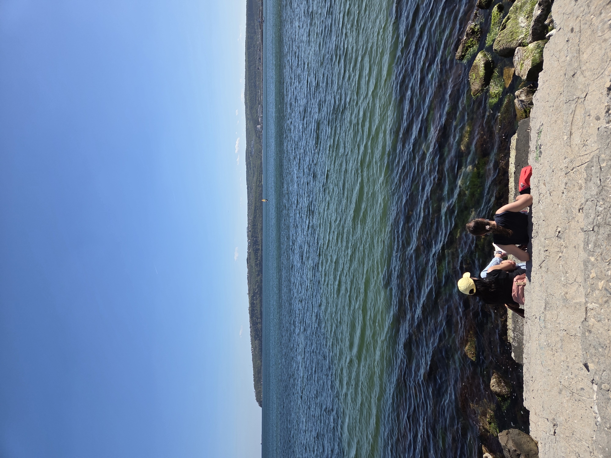 The image captures a view of the coast in Varna, Bulgaria. The composition is divided between the clear blue sky, the water stretching towards the horizon, and a concrete wall along the coastline. Two individuals are seated on the wall, partially visible. One is wearing a yellow cap and a dark shirt, the other a dark shirt. The water is a mix of blue and green, with visible waves, and rocky patches covered in algae near the wall. A strip of land is visible in the distance, marking the opposite side of the body of water.