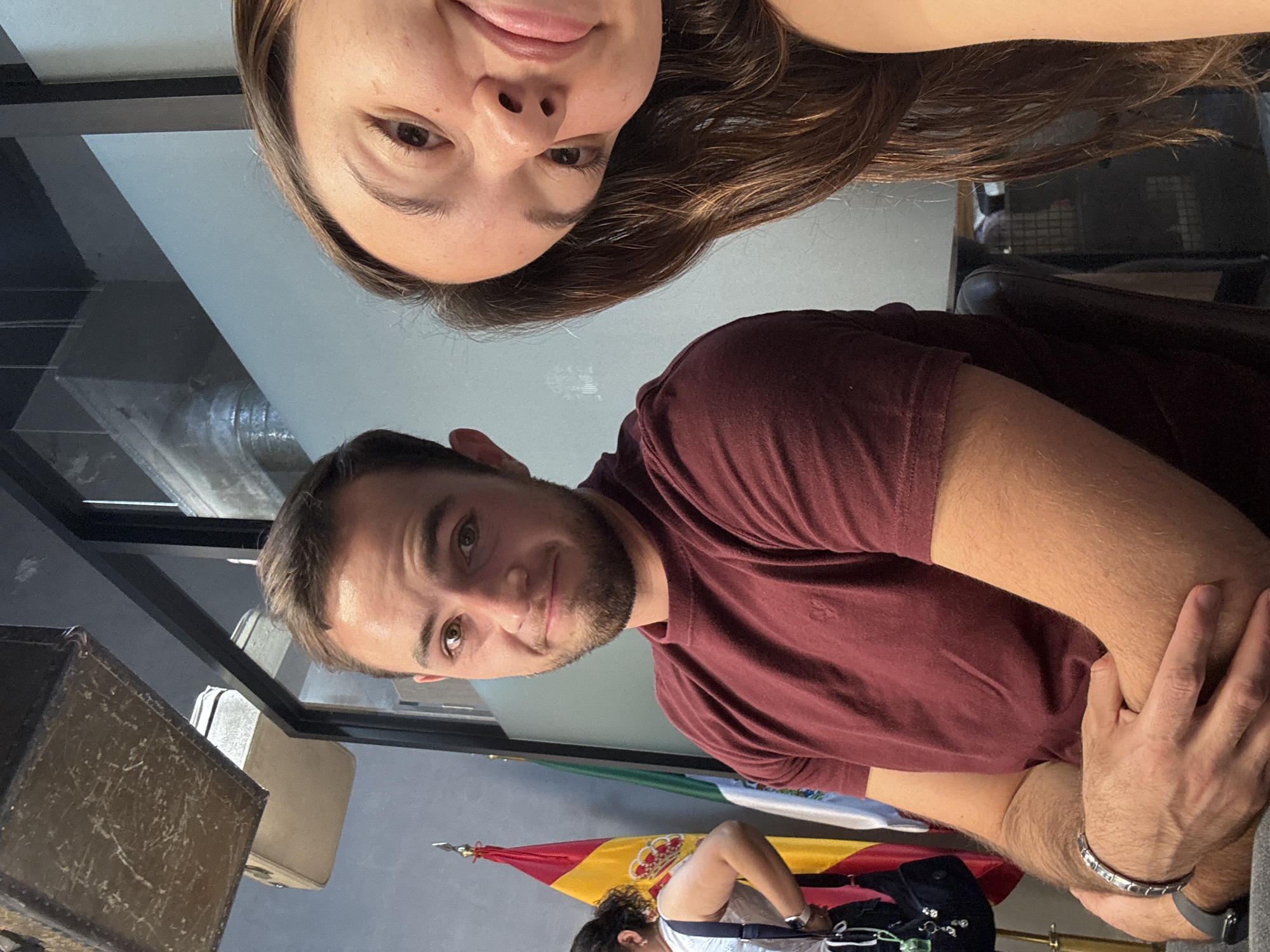 This image captures a close-up, high-angle selfie of two individuals, with a third person and various objects in the background, set indoors. The photo is rotated 90 degrees counter-clockwise.
In the foreground, a woman with long brown hair appears upside down in the top portion of the frame, smiling with her tongue slightly out, looking directly at the camera. Below her, occupying the mid-ground, is a man with short brown hair and a beard, wearing a maroon polo shirt. He is looking up towards the camera with a subtle smile, resting his left arm on something out of frame. On his left wrist, he wears a silver bracelet and a dark smartwatch.
The background reveals more of the setting. In the lower-left, a dark, textured box or vintage suitcase is visible. Behind it, what appears to be another lighter-colored suitcase can be seen. Further back and to the right, a Spanish flag with its red and yellow stripes and coat of arms is prominently displayed, partially obscured by a third person. This person is sitting, wearing a light-colored top and appears to be engaged in an activity, with a black bag and possibly some keys or a small trinket near them.
The setting appears to be an indoor space, possibly an office, school, or public building in Apetatitlán, Mexico, as suggested by the presence of the flag. The lighting is bright and even, indicating daytime. The expressions of the two main subjects convey a casual and playful mood. No visible text is discernible in the image.