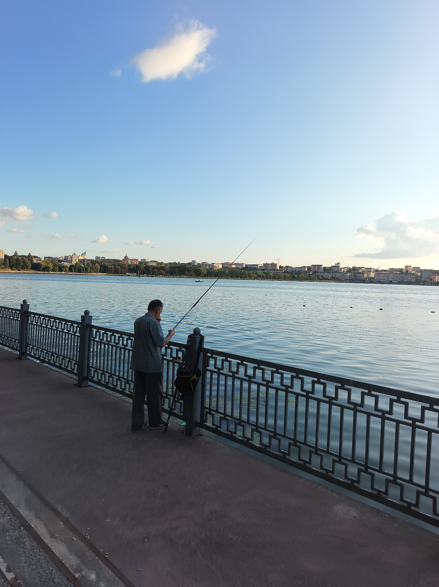 The image depicts a man fishing from the embankment of Ternopil Lake in Ternopil, Ukraine. The man, wearing a gray short-sleeved shirt and dark trousers, is holding a fishing rod that extends over the water. A black bag is hanging from a stand next to him. The lake is calm with gentle ripples, reflecting the clear blue sky which has a few scattered white clouds. Across the lake, a cityscape is visible with buildings, trees, and a flag. The embankment is paved with a dark surface and separated from the water by a decorative black metal railing. The overall scene suggests a peaceful, recreational moment on a sunny day.