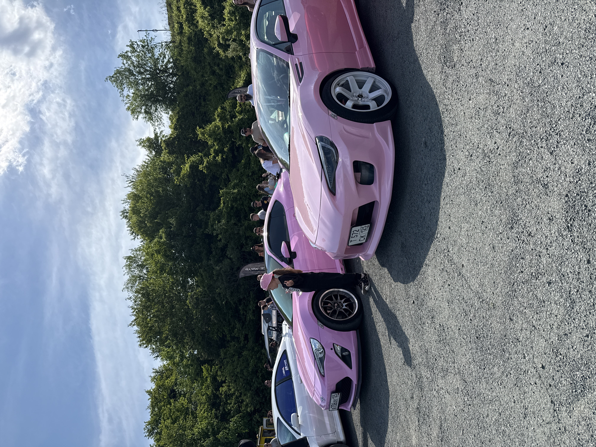 The image shows a line of parked cars, predominantly pink, at what appears to be an outdoor car meet. A young girl, wearing a pink beret and dark clothing, is standing near one of the cars. The scene is located outdoors on an asphalt-paved area, with trees and people visible in the background. License plates on the cars indicate that this photo was taken in Russia, in the city of Vladivostok. The vehicles appear to be modified sports cars.