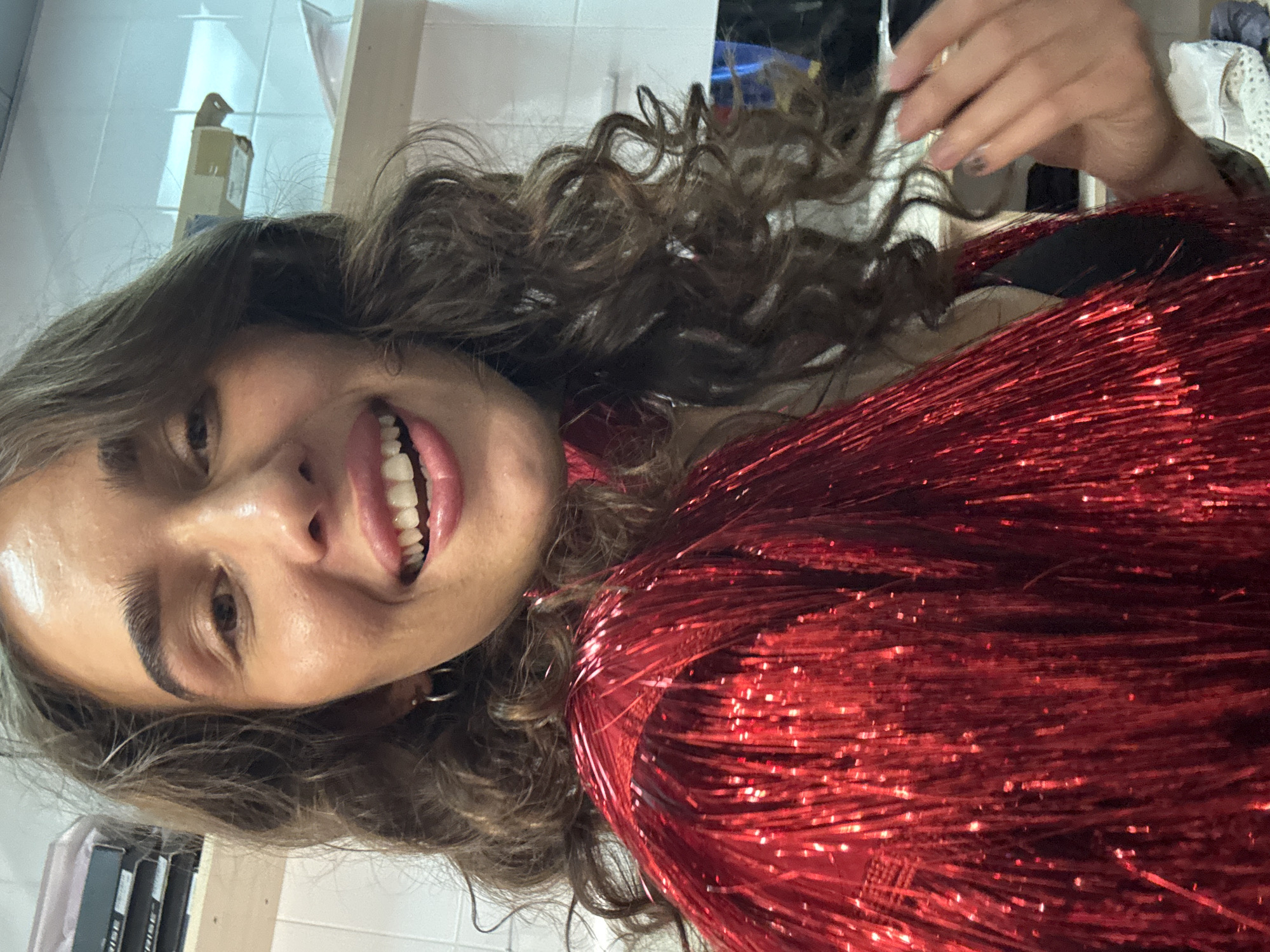 A female individual is captured in a close-up, selfie-style indoor photograph, displaying a wide smile with visible teeth. She has long, dark, wavy-to-curly hair that frames her face and shoulders. Her left ear (viewer's right) features a small silver hoop earring. She is wearing a prominent, voluminous garment or accessory made of bright, metallic red tinsel-like strips, which covers her chest and shoulders. A black garment is visible underneath the red tinsel on her right side (viewer's left). Her left hand (viewer's right) is slightly raised near her right shoulder, with fingers slightly splayed and short nails, some appearing to have dark polish or a design. The background indicates an indoor setting, with white tiled walls visible on the upper left and right. On the upper left, a stack of dark boxes labeled "RISE" is visible on a shelf. On the upper right, a light-colored object or shelf is partially obscured, and what appears to be a light fixture or vent is mounted on the wall. The lower right background shows indistinct objects, possibly a screen or mirror reflecting some blue light, and some white fabric or clothing. The image contains no visual information to confirm a location in Kazan, Russia.
