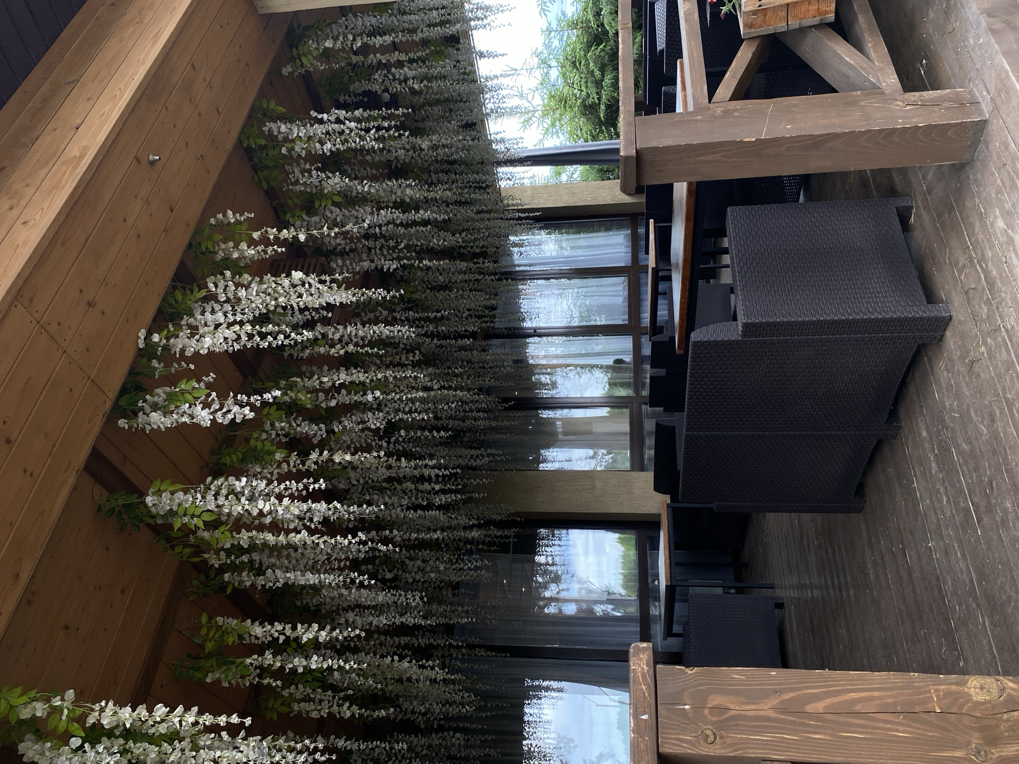 The image shows an outdoor seating area, possibly a veranda or patio, with a wooden floor and ceiling. Wicker chairs and tables are arranged on the floor. The area is decorated with long, hanging white flowers against a wall. Windows with sheer curtains are visible in the background. The location is likely a cafe or restaurant in Votkinsk, Russia.