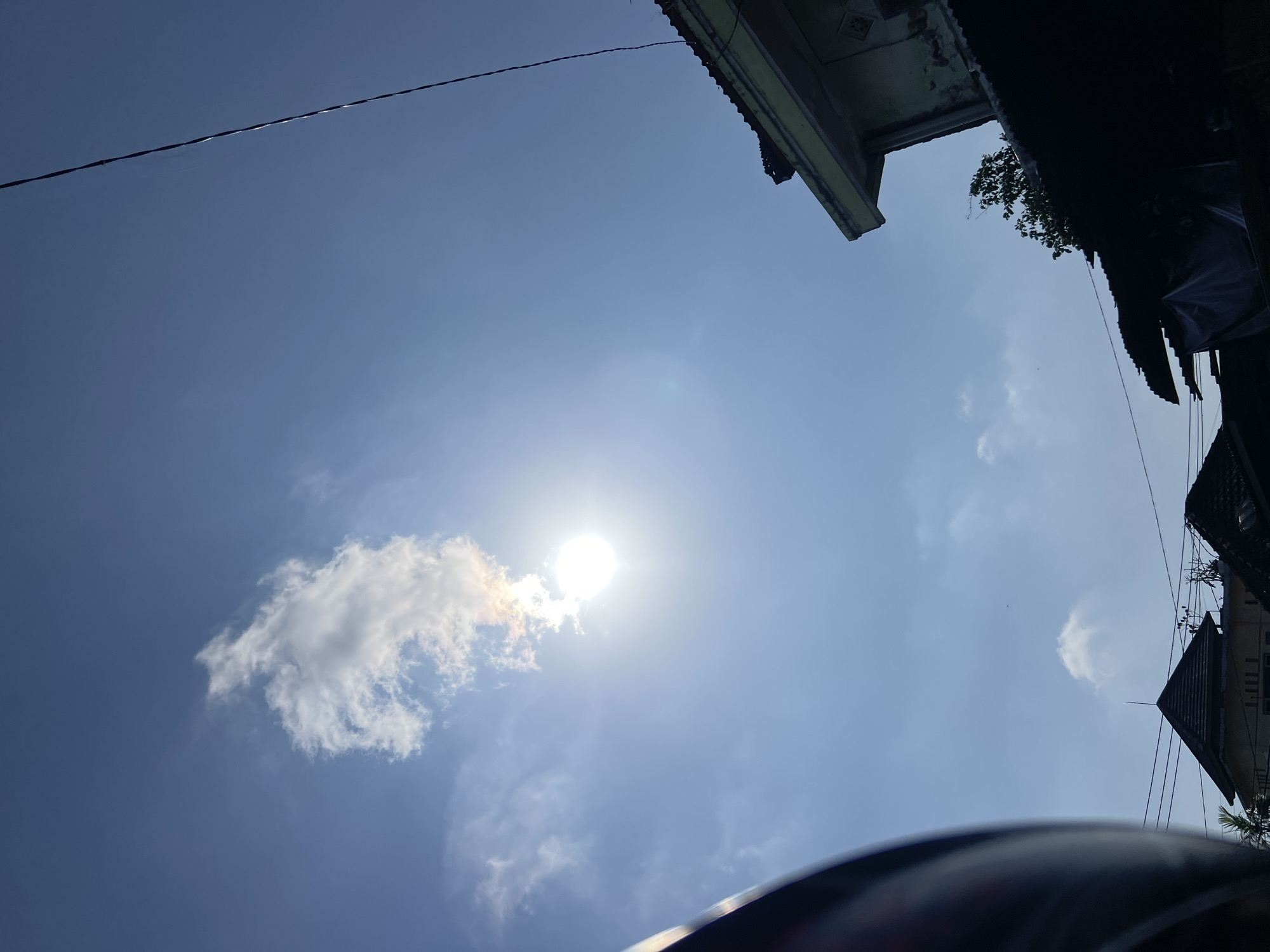 The image depicts a clear blue sky with a few scattered clouds in Denpasar, Indonesia. The sun is visible, shining brightly and creating lens flare. In the foreground, the edge of what appears to be a helmet or a curved, dark object is visible on the left side of the frame. On the right, the corner of a building with a light green facade and a tiled roof is present. Silhouetted rooftops of other buildings are visible at the bottom of the image, along with power lines crisscrossing the scene. A single dark electrical wire stretches diagonally across the upper right portion of the sky. The overall impression is of a bright, sunny day in an urban environment.