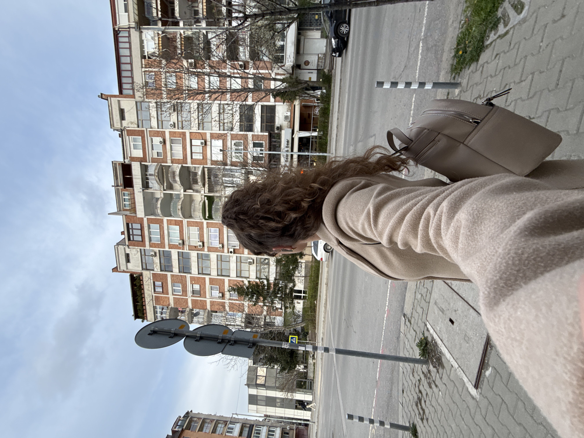 A medium shot shows a woman with curly brown hair and a beige coat and backpack on a sidewalk in Sofia, Bulgaria. The sidewalk is paved with gray bricks and has gray and white striped poles. Behind her, a street with sparse traffic is visible. Across the street, there is a multi-story building with red brick accents and white balconies. Street signs are visible on a pole near the sidewalk. The sky is overcast.