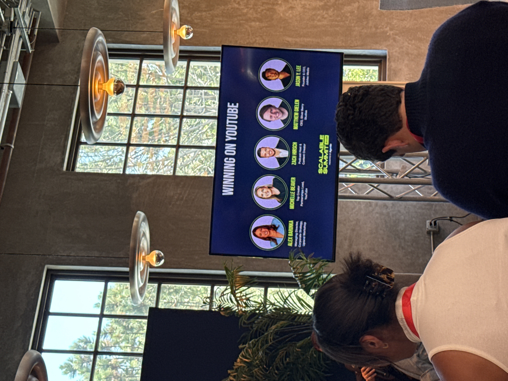 The image captures a presentation titled "Winning on YouTube" at the Scalable Summit in Los Angeles, California, United States. A screen displays headshots and names of panelists Alex Barinka, Michelle Beaver, Zach Hirsch, Matthew Gielen, and Jason Y. Lee, along with their titles and affiliations. Two individuals are partially visible in the foreground, one wearing a white shirt and the other a navy sweater, seemingly attending the presentation. The setting includes hanging light fixtures and windows with a view of trees, suggesting an indoor event venue.