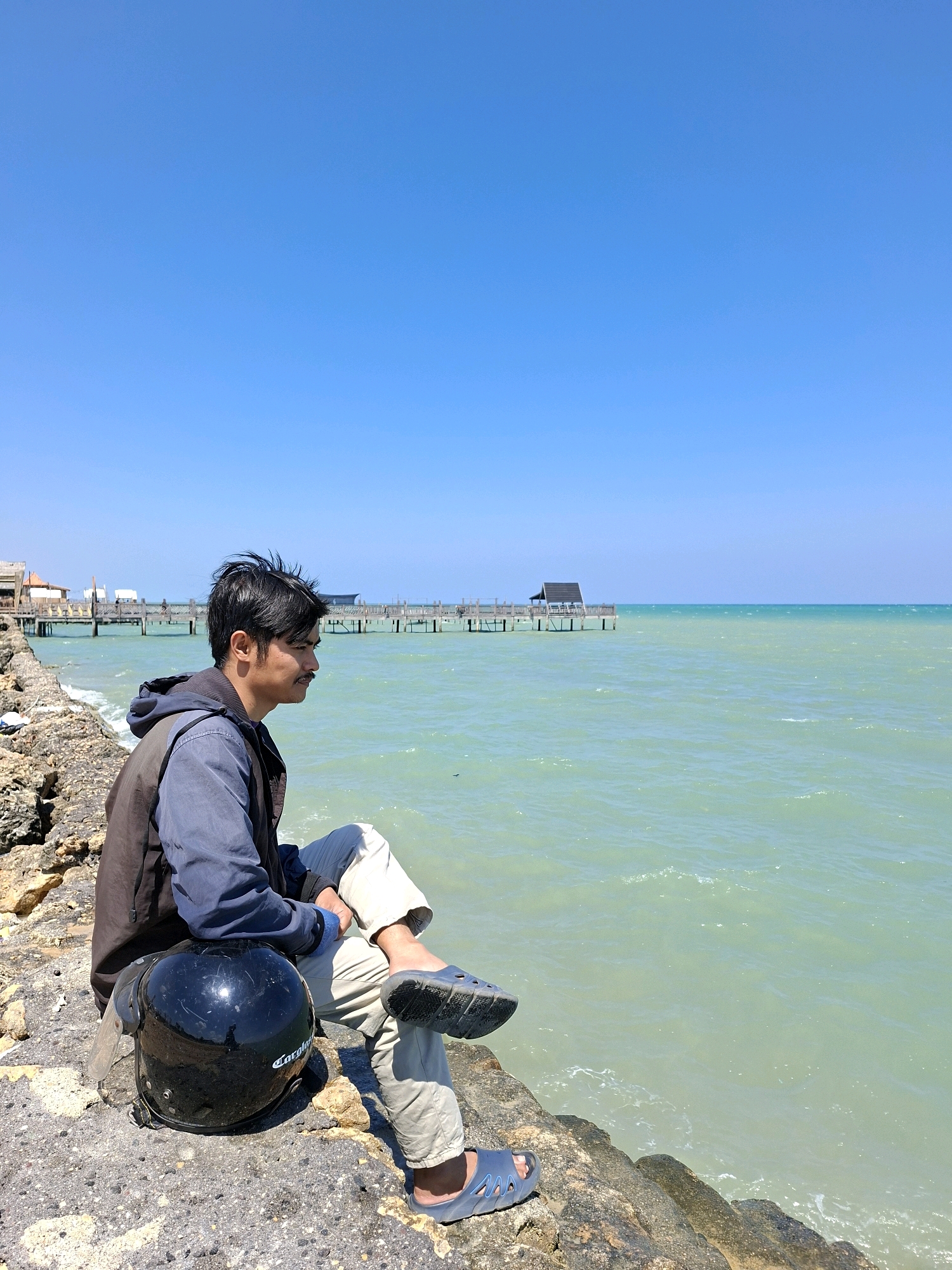 A man is sitting on a rocky shore in Sejati, Indonesia, looking out at the ocean. He is wearing a blue and black jacket, light-colored pants, and blue sandals. A black helmet is placed next to him on the rocks. The ocean is a light green color, and the sky is clear blue. In the background, there is a wooden pier extending into the water.