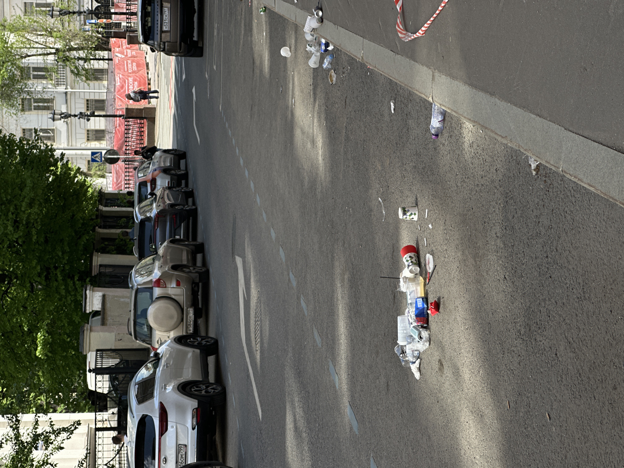 The image shows a street scene in Moscow, Russia. Several cars are parked along the left side of the street. There are several piles of trash scattered on the right side of the road and sidewalk. The day is sunny, casting shadows across the street. A pedestrian can be seen in the distance on the left side of the street behind some red banners. The street appears dirty and unkempt.