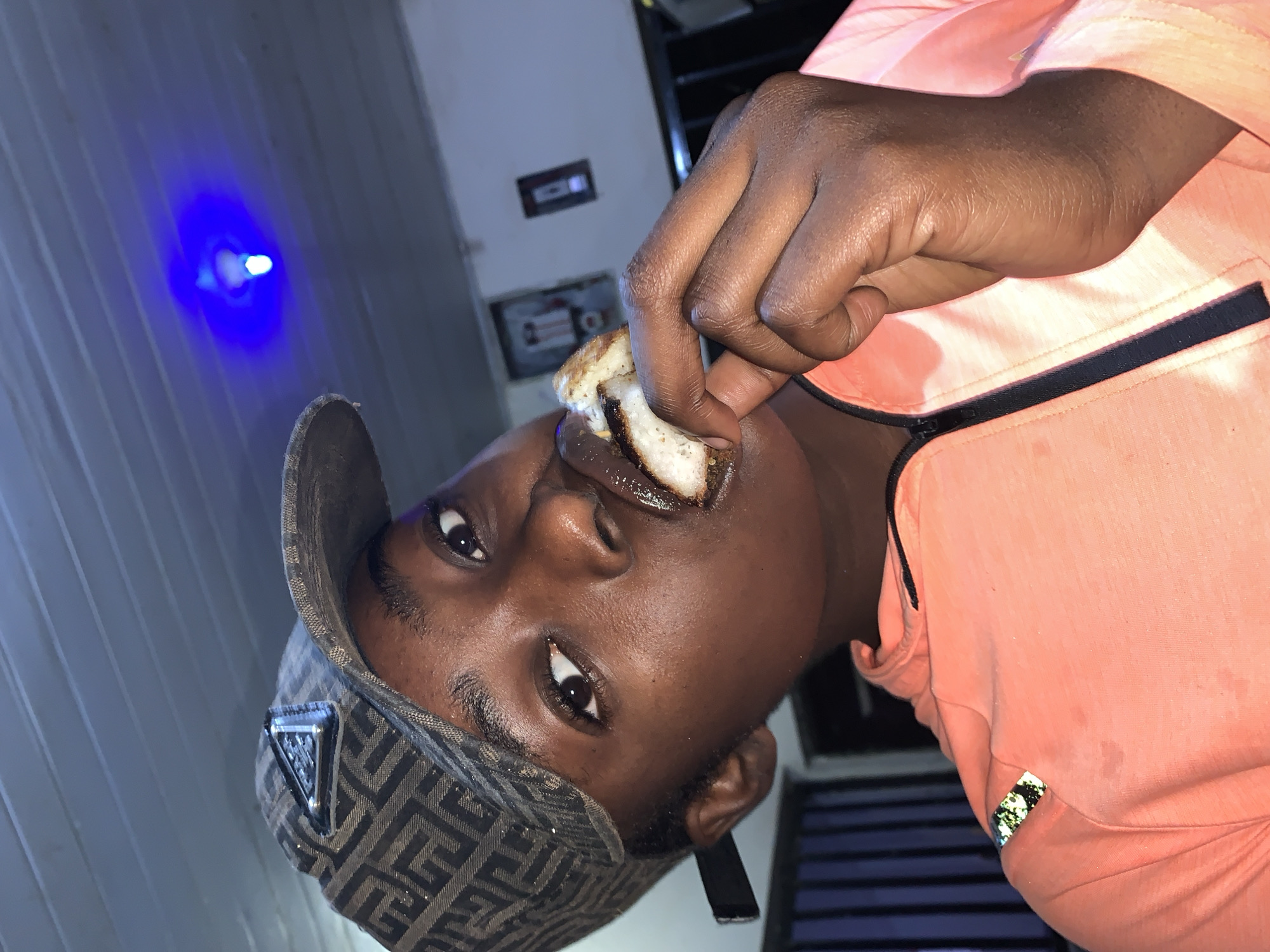 A young person with dark skin is eating a piece of toast. They are wearing a patterned cap and an orange athletic shirt with black trim. A blue light is visible in the upper left. The background suggests an indoor setting, possibly a room. There is no indication of the location being New Stadium, Nigeria.