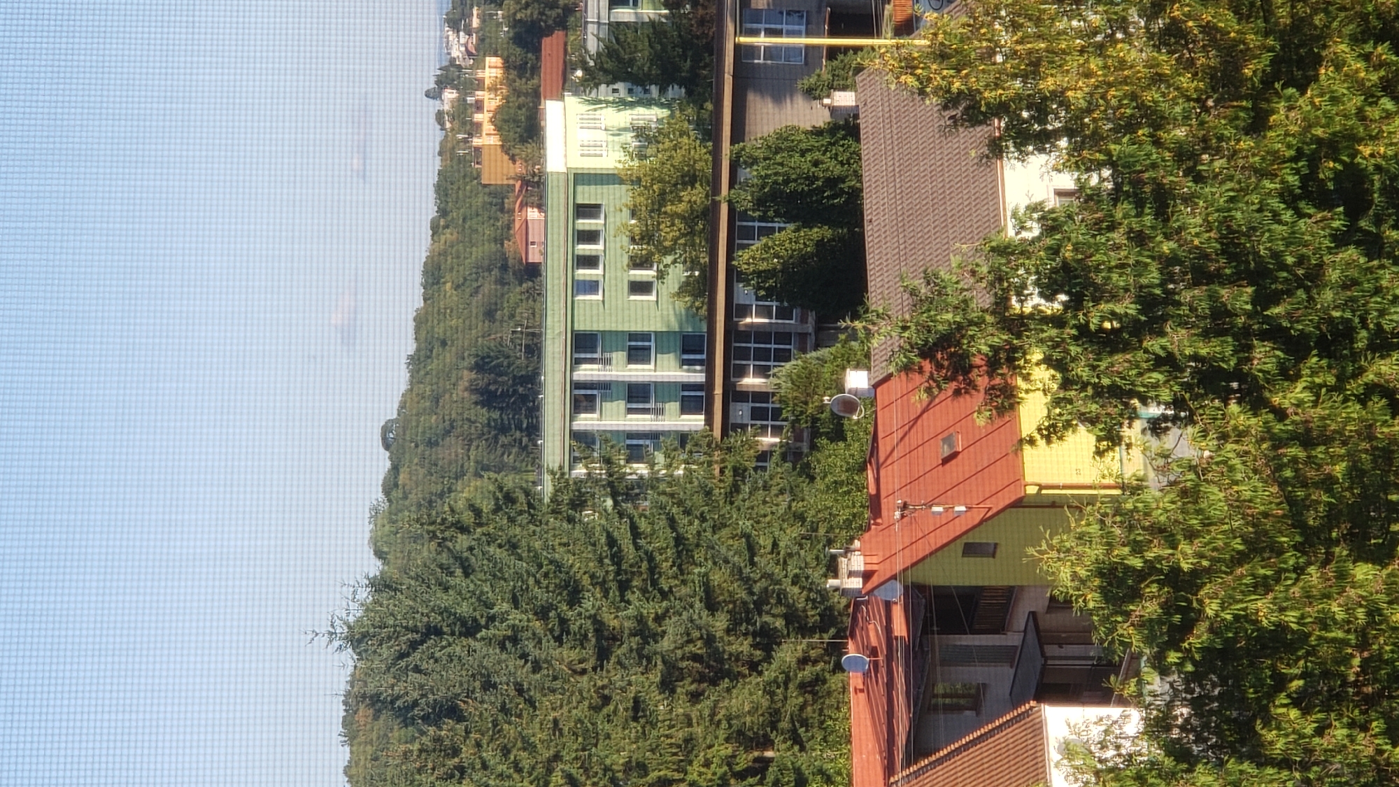 The image displays a view of a residential area with several buildings and trees, taken from an elevated position likely from a window with a screen. In the foreground, there are rooftops of houses with red and brown tiles. Behind these, a light green, multi-story building with many windows is visible, surrounded by dense evergreen trees. Further in the background, rolling hills covered in trees rise, with more distant buildings suggesting an urban or suburban landscape. The sky is clear blue with a few wispy clouds. The scene appears to be captured during daylight, likely in the late afternoon due to the lighting and long shadows. The presence of satellite dishes on some rooftops and the style of the buildings suggest a European setting. Specifically, this image was captured on the afternoon of September 17, 2023, at 15:48:59 in Prešov, Slovakia, at an altitude of 288 meters.