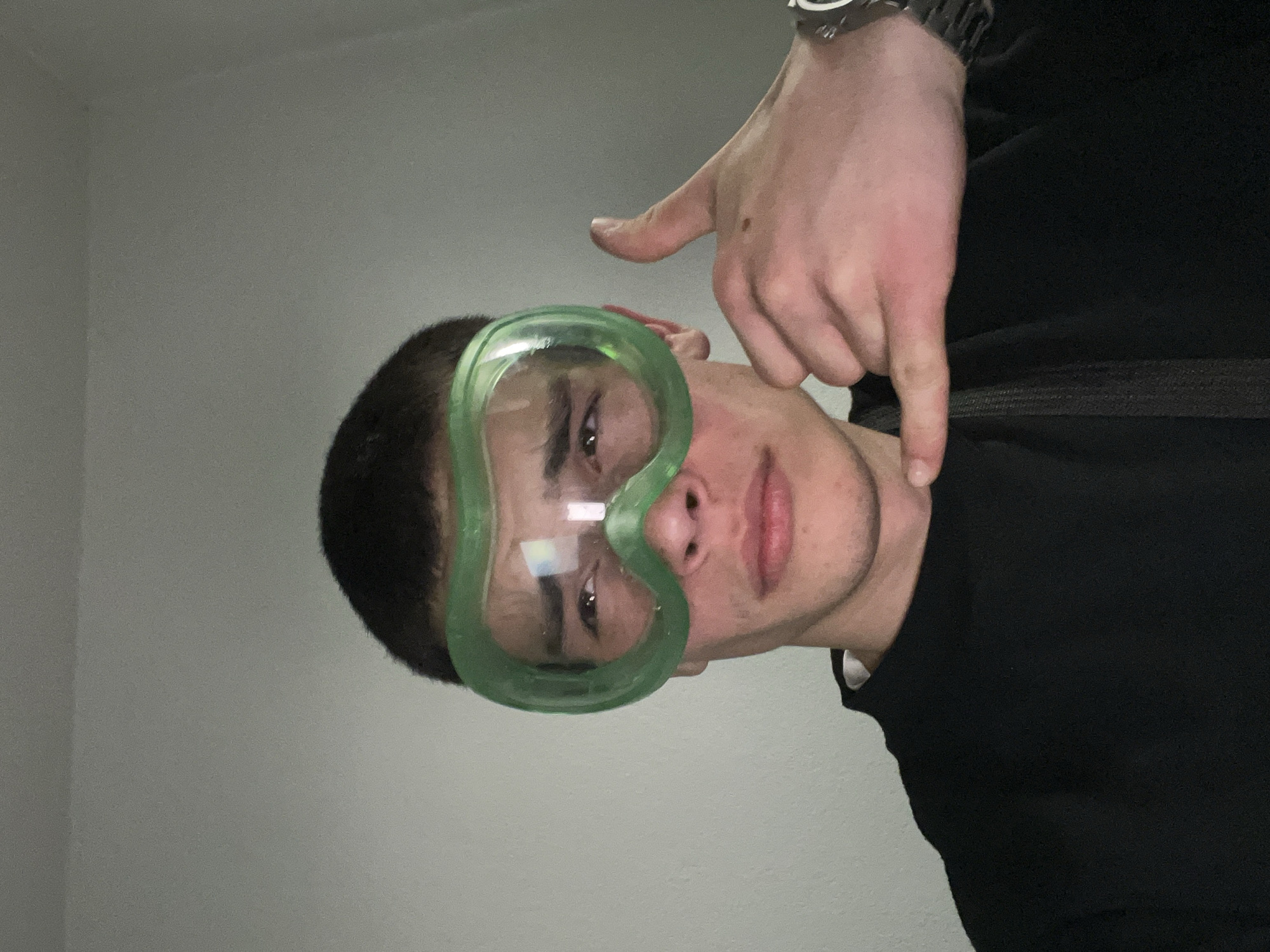 A young man with dark hair and light skin is wearing green goggles. He is pointing with his right index finger towards his collarbone. He is wearing a black shirt. A watch is visible on his left wrist. The background appears to be a plain, light-colored wall. The photo's origin, Karavelovo, Bulgaria, cannot be verified from the image.