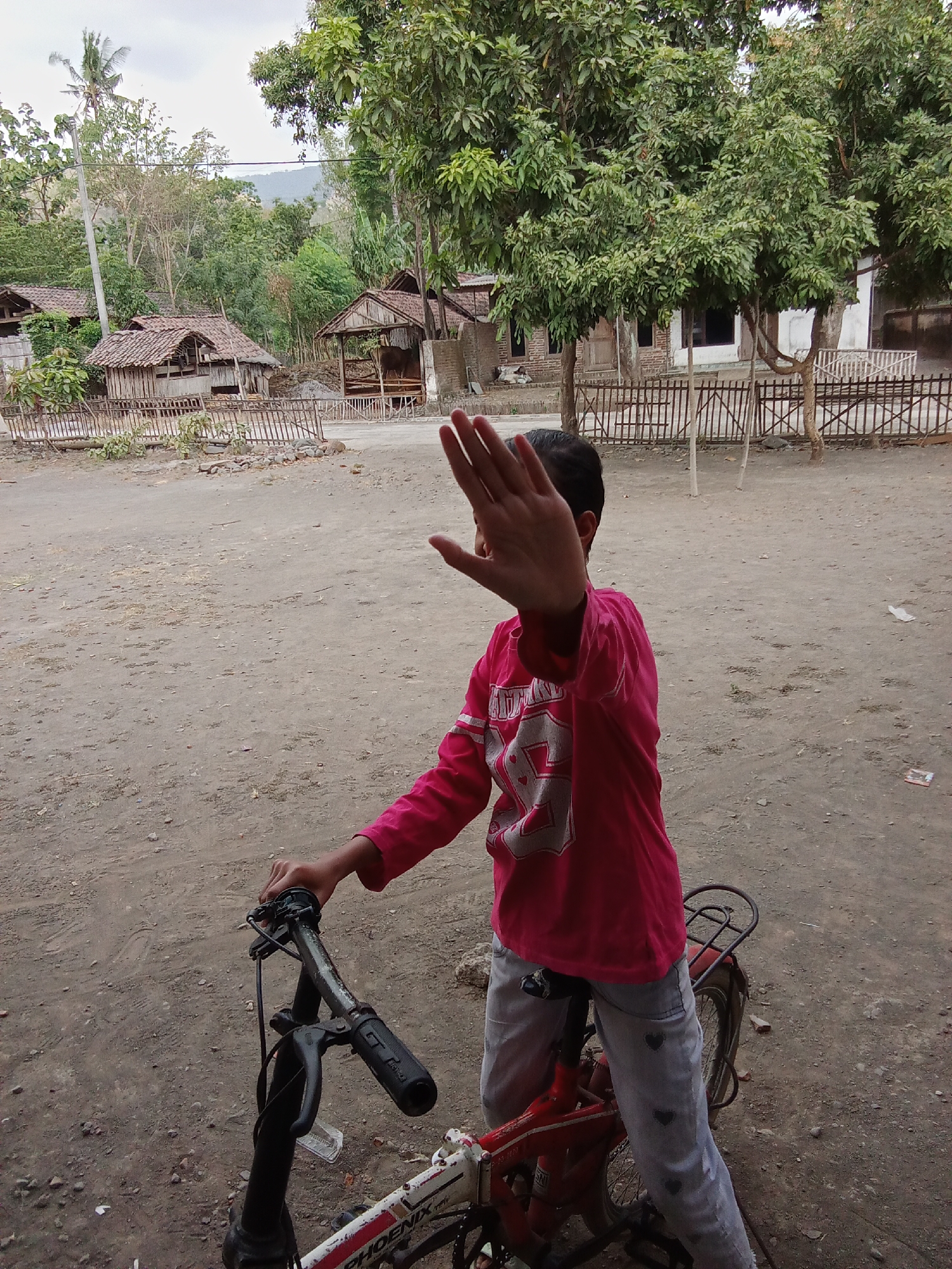 In the city of Selowogo, Indonesia, a child on a bicycle blocks the camera with their hand. The child wears a bright pink shirt with white lettering and light gray pants adorned with dark gray heart shapes. They are positioned on a bicycle with a red frame, white accents, and a black seat. The scene unfolds on a dirt ground, likely a pathway or open area, with a rustic wooden fence in the background. Behind the fence, several small, traditional wooden houses with tiled roofs are visible, nestled amongst lush green trees and vegetation. The overall atmosphere suggests a rural or semi-rural setting.