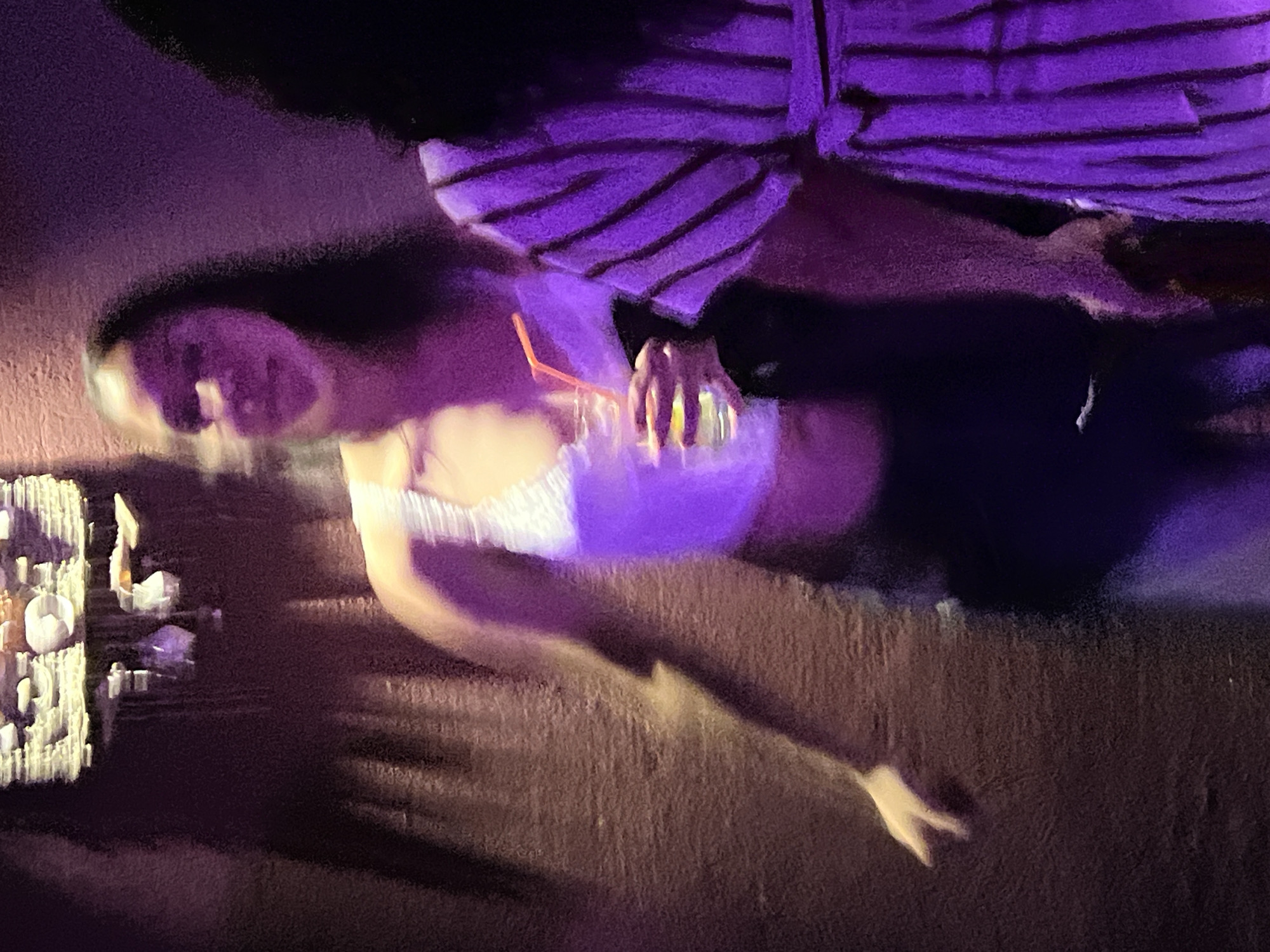 The image shows two individuals at night, dimly lit with purple and yellow lighting, possibly in a bar or club. The person in the foreground, on the left, is a woman with long dark hair, wearing a white sparkly top and dark pants. She is holding a clear glass with a red straw, filled with a light-colored beverage. Her facial features are blurred due to motion, but she appears to be looking forward. To her right, another person is partially visible, wearing a dark garment with vertical purple and white stripes, and a belt. The background consists of a textured, reddish-brown wall with a shadow cast on it. Above and slightly to the left, a decorative light fixture hangs from the ceiling, also illuminated with warm yellow light, and there are some hanging decorative elements. The overall impression is one of a lively, possibly social, indoor setting. The prompt mentions Herceg Novi, Montenegro, but there is no visual information in the image to confirm this specific location.