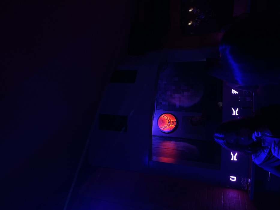 The image depicts a dimly lit indoor scene, saturated with blue and purple ambient lighting, suggesting an evening or nighttime setting. Two individuals are partially visible on the right side, their heads and upper bodies illuminated by the blue light, indicating they are actively engaged with the surroundings.
A central feature is a circular, orange-red illuminated fixture embedded in a dark panel. Within this light, the distinctive Jägermeister logo—a stag's head with a cross between its antlers—is clearly visible. Above and behind this panel, a dark surface with a subtle checkered or tiled pattern can be discerned.
Adjacent to the Jägermeister fixture, on a vertical panel, several white or light blue illuminated symbols or letters are displayed. These symbols, from top to bottom, resemble 'F', 'Y', 'Y', and 'D'. The floor in the foreground and to the right appears to be made of wood, reflecting the blue light. The overall atmosphere and setup suggest a recreational area, possibly a bar or themed entertainment space, located in Drapetsona, Greece, where people are interacting with illuminated displays.