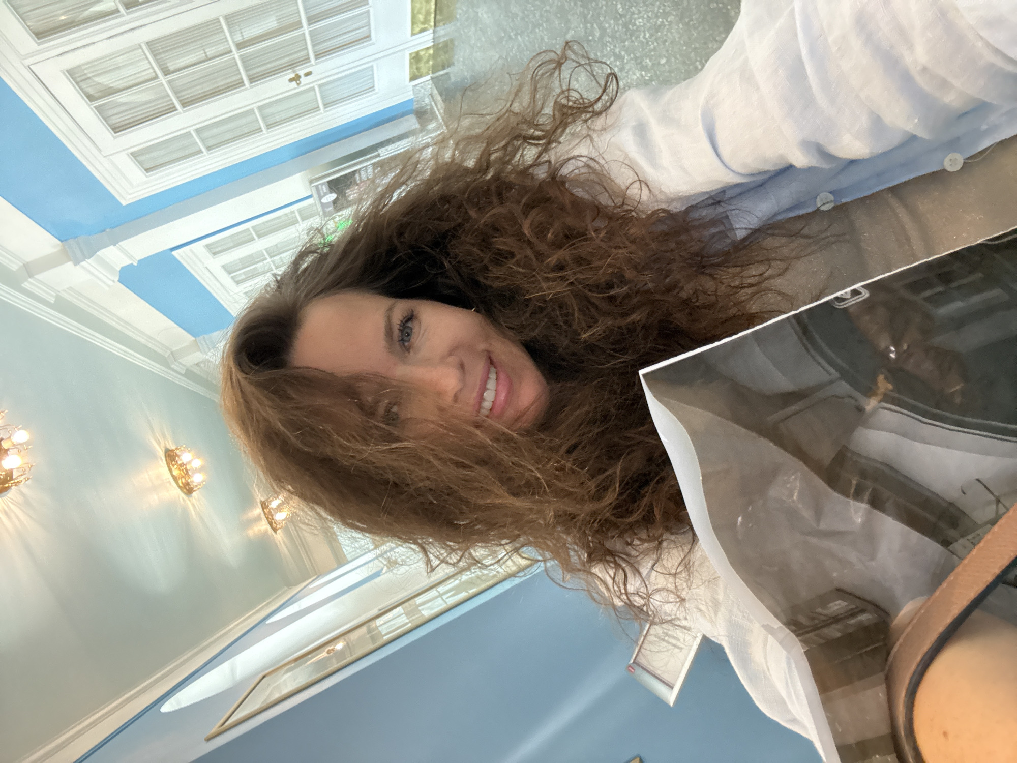 The image shows a woman with long, curly brown hair, smiling at the camera. She is wearing a light blue linen shirt. She is holding a flat, rectangular object, possibly a brochure or a magazine, in front of her. The background appears to be an interior hallway with light blue walls, white trim, and ornate ceiling lights. There are white double doors visible in the background. The flooring is a reflective, light-colored material. The woman's location is not specified in the image or its context.