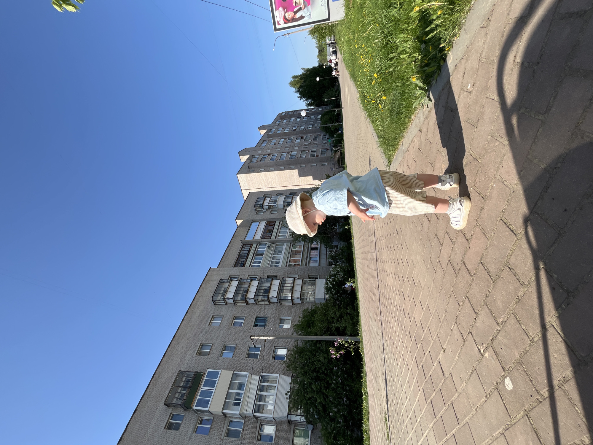 A low-angle shot depicts a child standing on a brick sidewalk in what appears to be the city of Chaikovsky, Russia, indicated by the presence of a typical Soviet-era apartment building in the background. The child, wearing a light-colored hat, a light blue shirt, and striped pants paired with white sneakers, is positioned slightly off-center in the frame. The scene is bathed in sunlight, casting a shadow of the child onto the sidewalk. The sidewalk abuts a grassy verge with yellow flowers. A multi-story grey brick apartment building with multiple balconies is seen in the background, extending into a clear blue sky. A billboard is partially visible above the apartment building. The overall impression is one of a sunny day in a residential area.