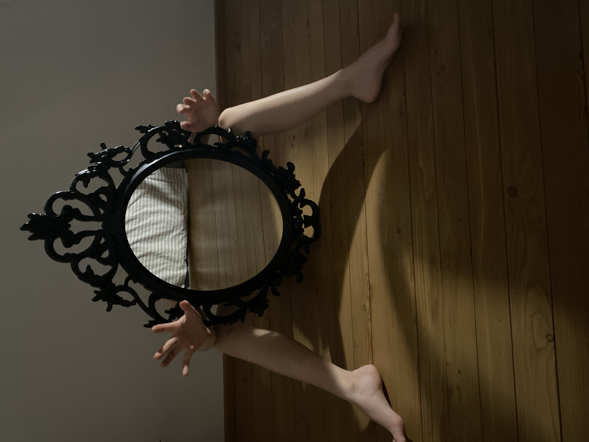 A slightly canted, medium-range indoor shot depicts an ornate black oval mirror with body parts seemingly emerging from within. The mirror, positioned at an angle, reflects a striped bedding surface on what appears to be a bed. Coming out of the top of the mirror are two limbs: an arm ending in a hand with outstretched fingers. Exiting from the bottom of the mirror is another arm ending in an open hand. These limbs appear to connect to two legs that extend to the right onto a vertical, wood-paneled surface, likely a wall. The feet on the ends of these legs are bare. The wall behind the mirror is a light gray-white, and the overall lighting suggests an artificially illuminated setting. There is no information to suggest the location is the city of Chaikovsky, Russia.