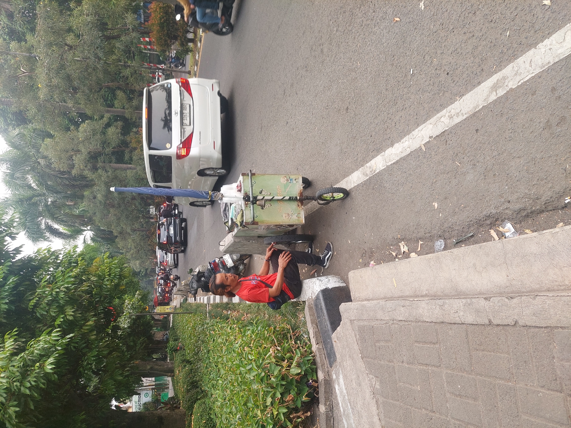 A man in a red t-shirt and black pants sits on a concrete curb next to a street in Bogor, Indonesia. He has light brown skin, dark hair pulled back in a ponytail, and is wearing black sneakers. He is looking to his right. Beside him is a green cart with two wheels and a blue umbrella. The cart has a bell attached to it and an image of cartoon children on the side. A white van and several motorcycles are visible on the road behind him, indicating traffic. Trees line the left side of the street, and a concrete sidewalk is visible in the foreground. The street appears to be paved with asphalt and has a white line marking the lane.
