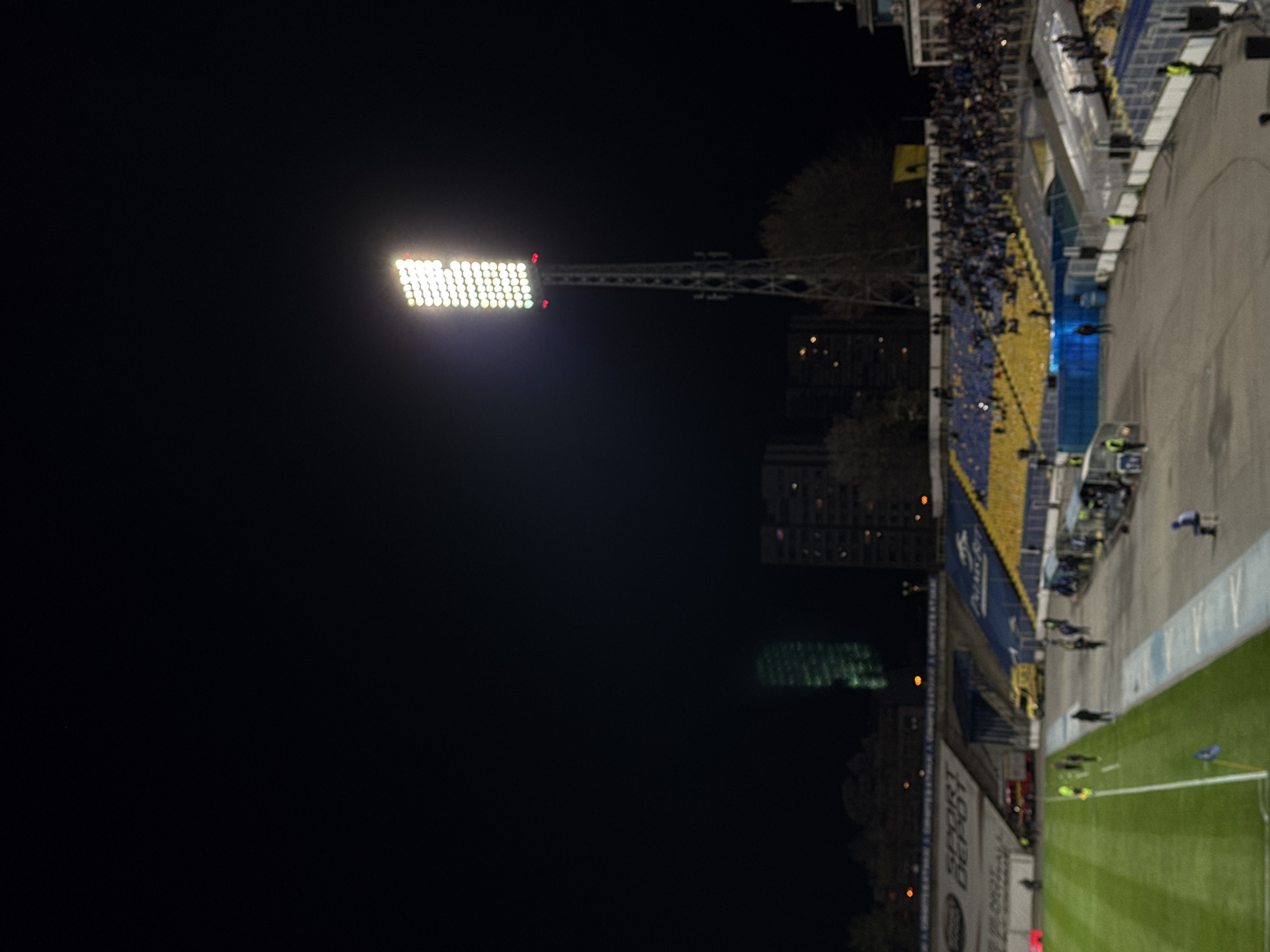 The image shows a night view of a football stadium. The stadium is illuminated by bright floodlights. A section of the stands is filled with spectators. In the background, buildings can be seen. The stadium is the Vasil Levski National Stadium in Sofia, Bulgaria.