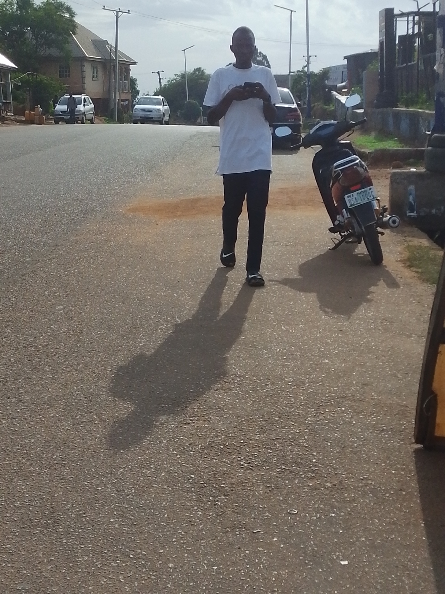 The image depicts a street scene in Jos, Nigeria. A man in a white t-shirt and dark pants is walking on a paved road while looking at his phone. Behind him, a black motorcycle is parked, with a visible license plate that reads "BSA 109QC." The road stretches into the distance, where two cars are parked; one silver and the other appears white. Buildings and trees are visible on either side of the road. The sky is overcast. The man's shadow is cast on the road in front of him.
