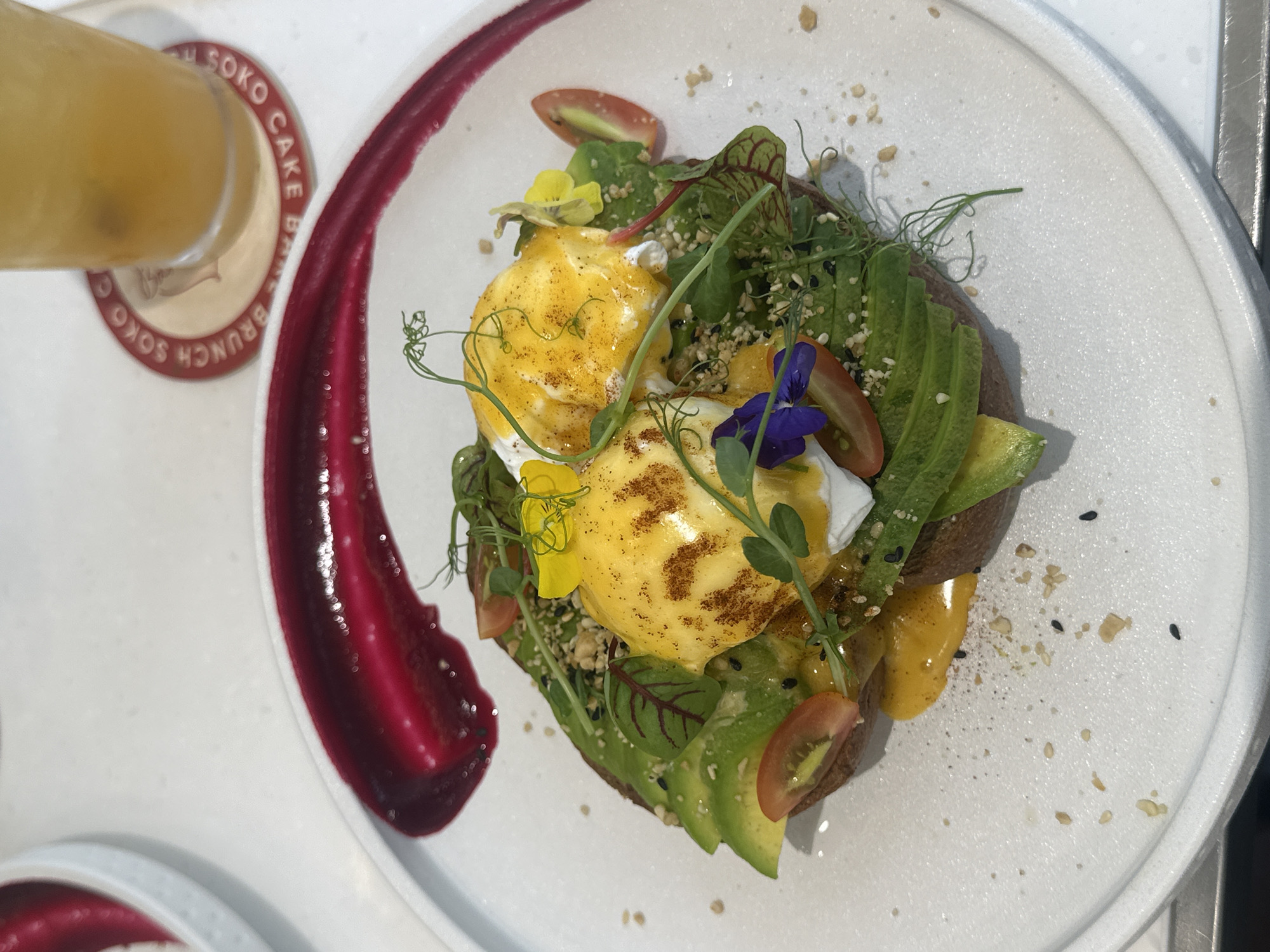 The image displays a brunch dish consisting of avocado toast with two poached eggs, presented on a white, textured ceramic plate. The dark-colored toast base is topped with fanned slices of green avocado, green microgreens, small red-veined leaves, and halved red cherry tomatoes. Two poached eggs, each covered in a bright yellow sauce with light brown patterned dusting, are centered on the toast. The dish is garnished with green pea shoots, white sesame seeds, black sesame seeds, finely chopped nuts, one yellow edible flower, and one purple edible flower. A curved smear of vibrant magenta sauce is present on the upper right portion of the plate. The plate rests on a white tabletop. In the upper right background, a clear glass containing a yellowish beverage sits on a round, red-rimmed coaster bearing the text "SOKO CAKE BRUNCH". A partial view of another white dish is visible in the upper left background. The location is Soko Cake Brunch, specifically identified by the coaster, which has a known establishment at 62 Võ Văn Ngân, Bình Thọ, Thủ Đức, Thành phố Hồ Chí Minh, Vietnam.