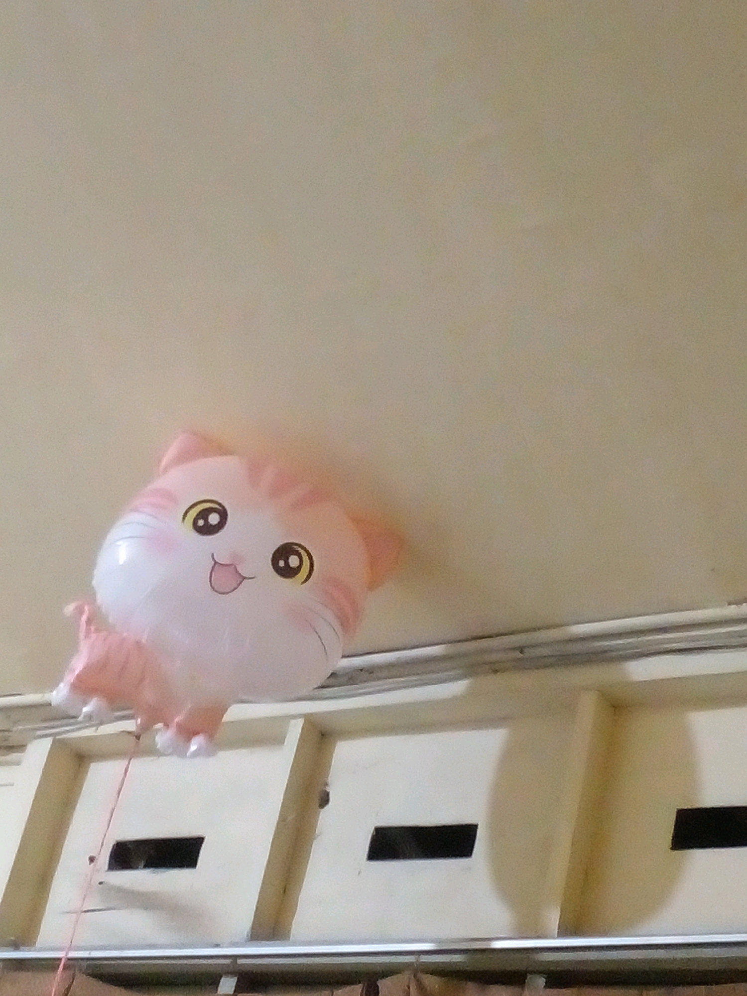 An inflated, light pink and white balloon shaped like a cartoon cat is suspended from the ceiling. The balloon has large, round, yellow-rimmed eyes with black pupils and a smiling mouth with a pink tongue. Pink stripes adorn the top of its head and its body. The balloon appears to be a party decoration. The background is a neutral-colored ceiling and a section of what looks like wall paneling with rectangular openings. A thin red string is attached to the balloon and hangs down. The image was captured in Pungangan, Indonesia.