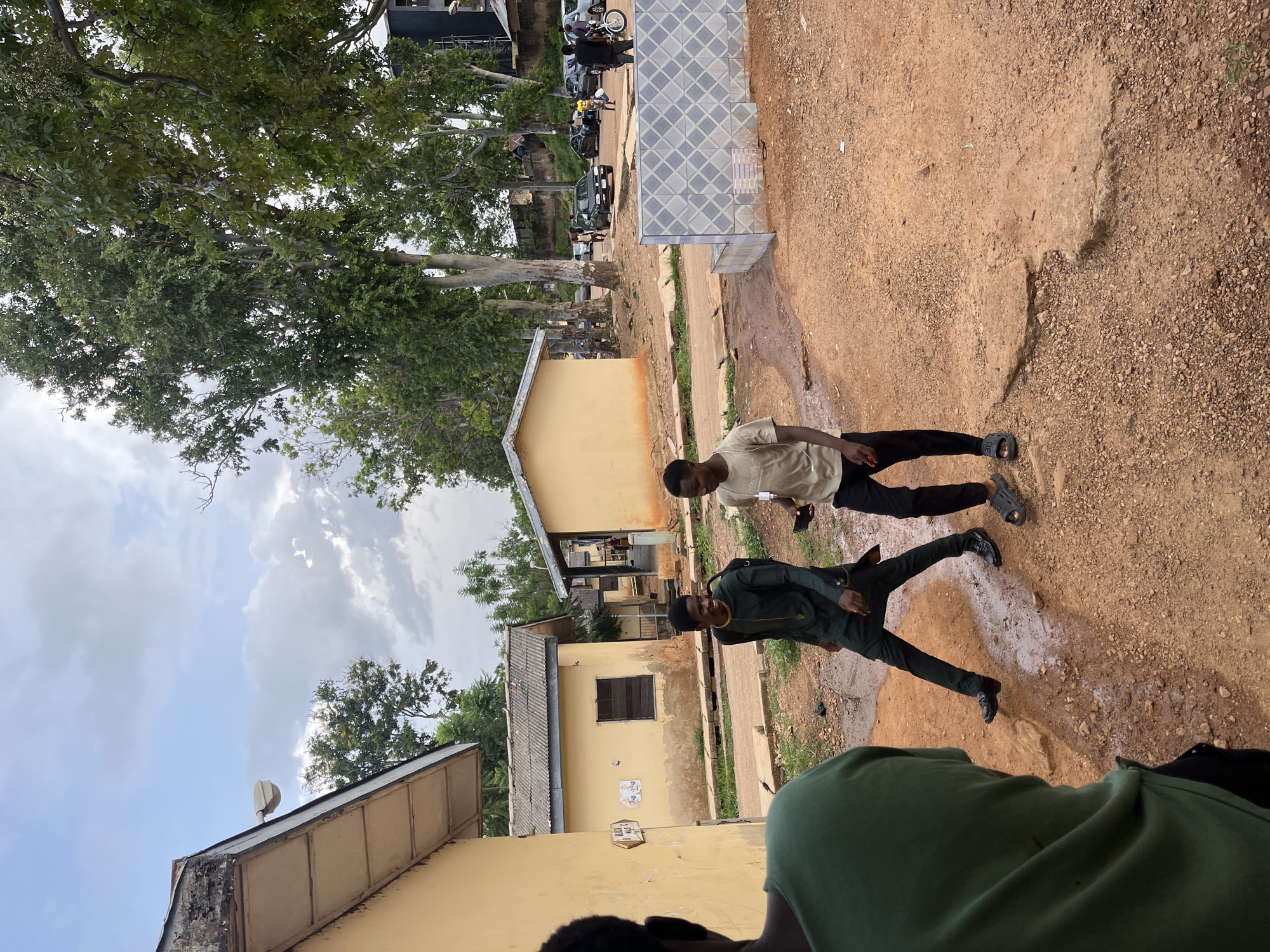 The image depicts an outdoor scene in Jos, Nigeria, with a focus on two young men walking on a dirt path. The path is partially wet and muddy, with some scattered stones and patches of dry earth. To the left, a person wearing a green t-shirt is partially visible from behind, facing away from the camera.
In the foreground, two men are walking towards the right. The man on the left is wearing a dark green traditional garment with gold embroidery and black shoes. He has a black backpack slung over his left shoulder. The man on the right is wearing a light-colored t-shirt, black pants, and dark sandals. He is holding a dark object, possibly a phone, in his right hand. Both men appear to be in motion and are looking in the same direction, to the right of the frame.
In the background, there are several low, single-story buildings with yellow painted walls and corrugated metal roofs. One building has a small window with a dark frame. There is a signpost on the left wall of one building, and some posters are affixed to the wall. Further in the background, a row of tall trees with lush green foliage lines the area. Beyond the trees, several cars are parked, and more buildings are visible. A short, tiled wall with a diamond pattern is present in the mid-ground to the right. The sky is overcast with patches of blue visible. The overall environment appears to be a residential or institutional compound.