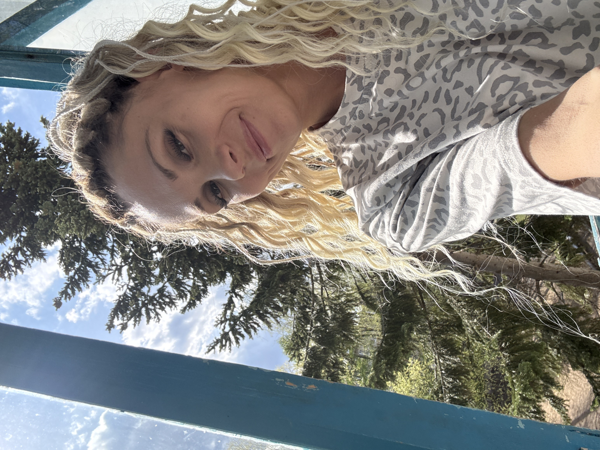 A blonde woman with curly hair and a braided section is looking at the camera from an elevated position. She is wearing a light gray patterned shirt. Below her, trees and the sky are visible through what appears to be a blue-painted frame or window. The location is likely Rusanovo, Russia, given the request.