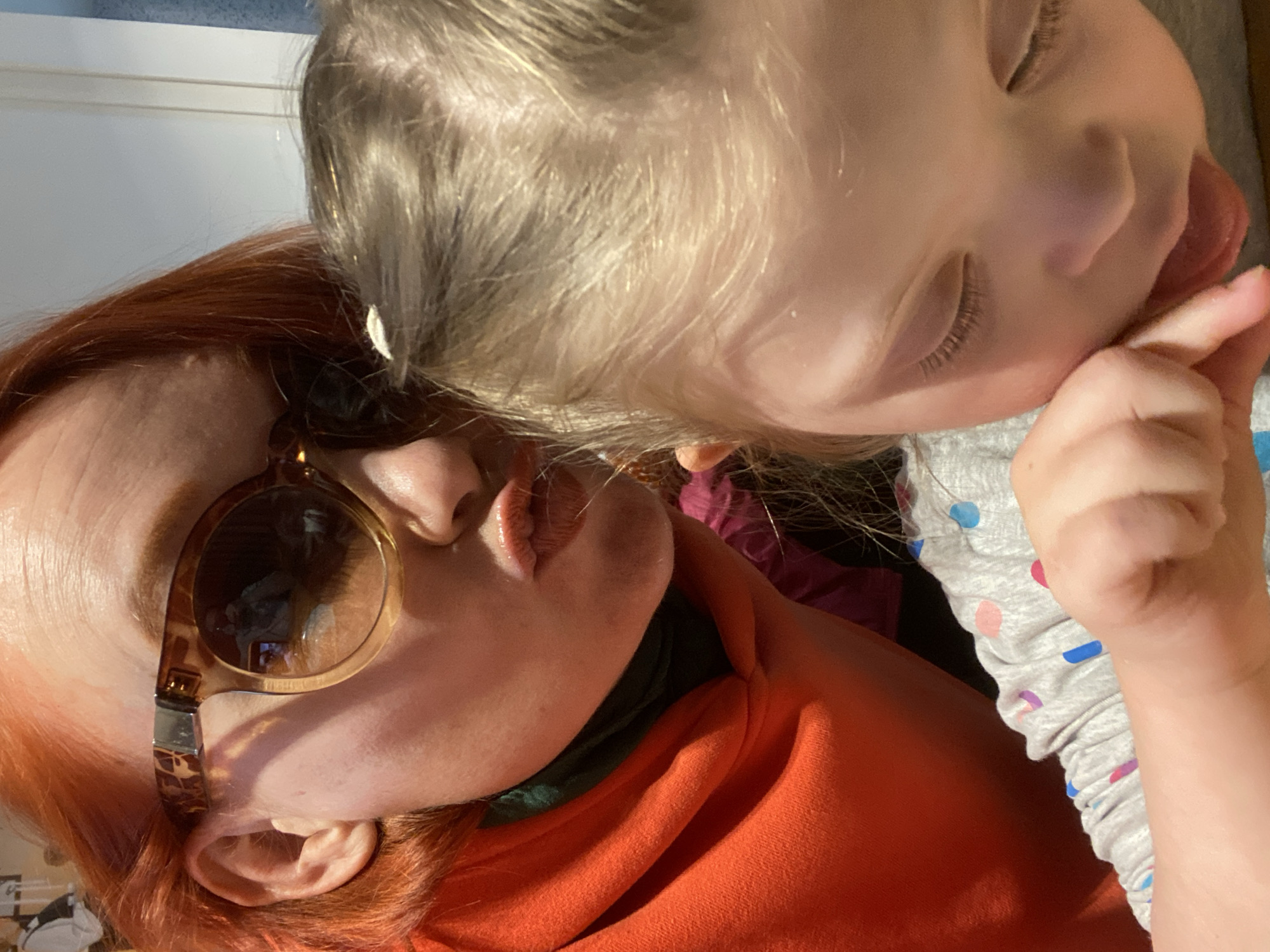 A close-up indoor shot depicts an adult woman with reddish-orange hair wearing round, tortoiseshell-rimmed glasses, lying next to a young child with blond hair. The child is mostly out of focus and facing the camera. The child has its eyes closed and is wearing a gray, patterned shirt. The adult woman is wearing an orange shirt with a green collar. The image is rotated so that the subjects are oriented on a diagonal. The location cannot be determined.