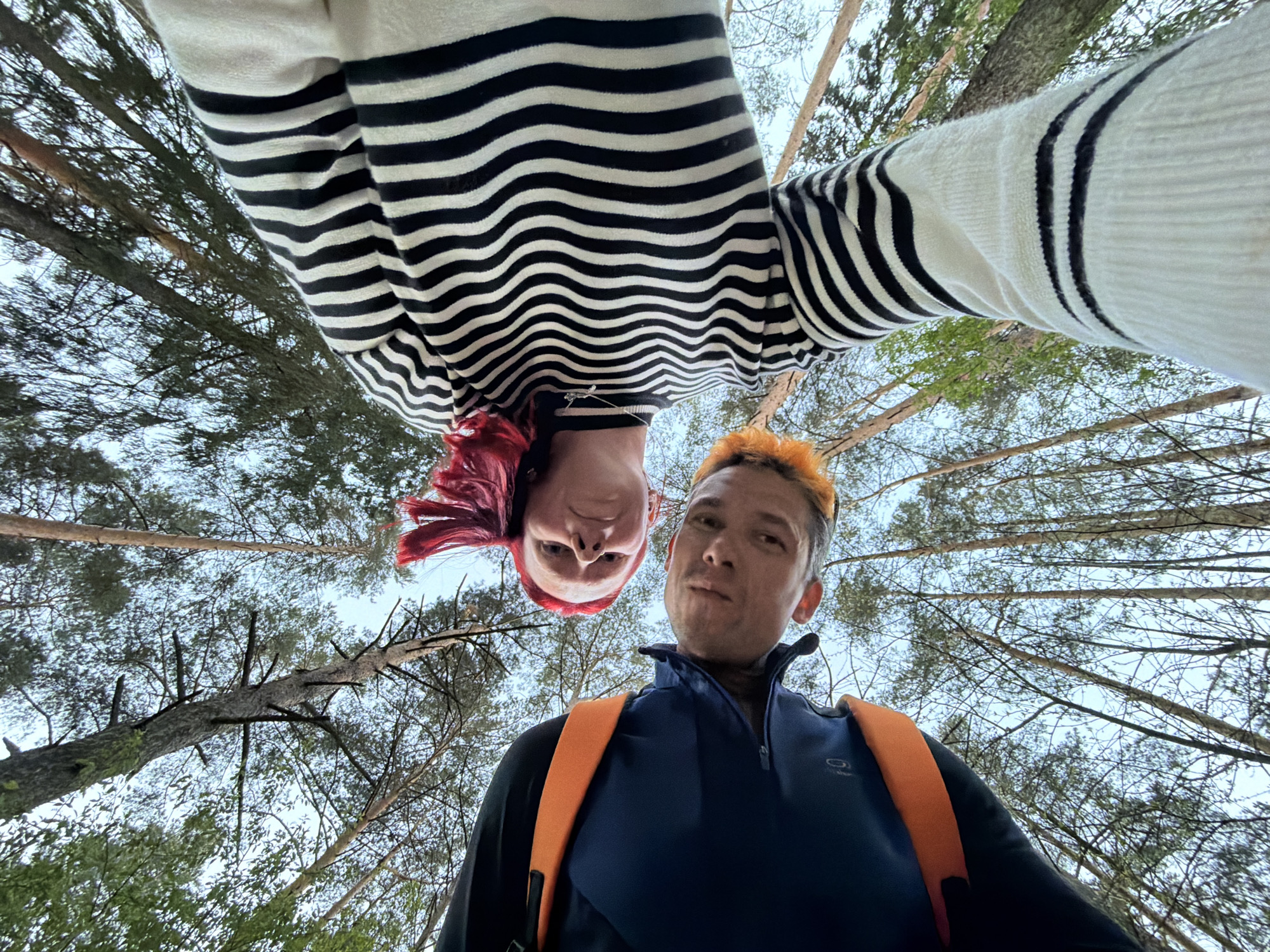 The image depicts two people standing in a forest area in Chaikovsky, Russia. The photograph is taken from a low angle, looking up at the subjects. One person, with dyed red hair, is seen wearing a black and white striped sweater. The second person has dyed orange hair and is wearing a blue zip-up jacket with orange backpack straps. The background shows tall trees reaching towards the sky.