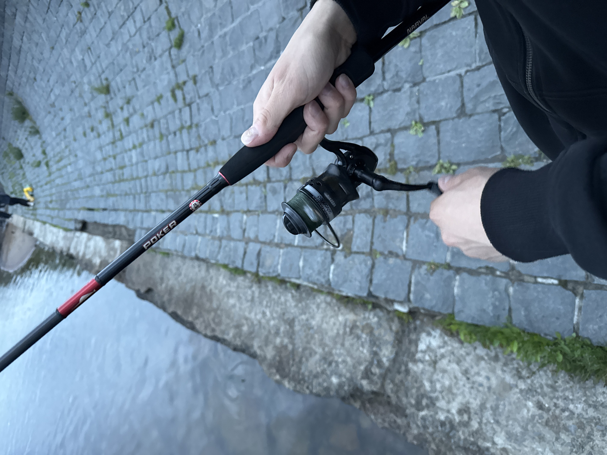 The image depicts a person holding a fishing rod and reel near a water body. The ground is paved with cobblestones, and there is a concrete embankment along the water's edge. In the background, other people are visible, one of whom is wearing yellow shoes. The fishing rod has the brand name "POKER" visible on it, and the reel has "dovoked" and "60331" printed on it. The overall setting appears to be an urban environment, possibly a canal or riverbank in the city of Saint Petersburg, Russia, given the architectural style of the paving. The lighting suggests it is either early morning or late afternoon.