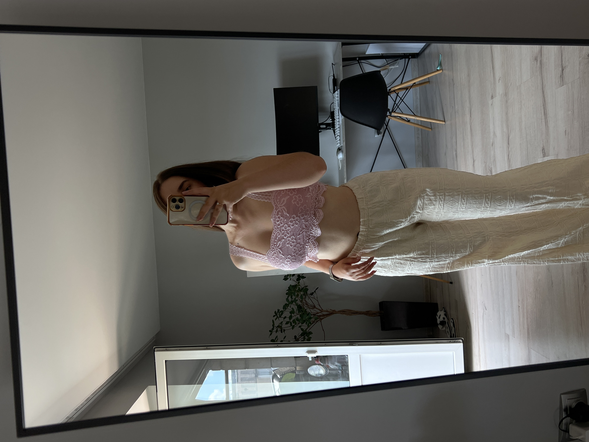 A person is taking a mirror selfie in a room. The person is wearing a light pink lace bralette and cream-colored wide-leg pants. The room appears to be a bedroom or living area, with a large mirror, a desk with a computer monitor and a chair, a tall white cabinet, and a potted plant. The flooring is light-colored wood. The image does not contain any specific information about the location being Ternopil, Ukraine.