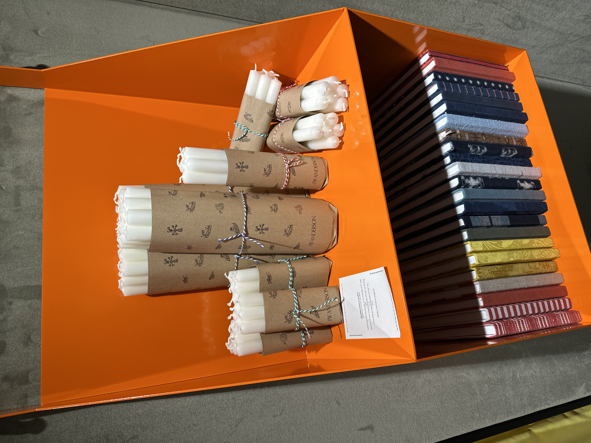 This media file captures an indoor retail display or product presentation, likely in London, United Kingdom. The central subject is a large, glossy, bright orange container with angled sides, possibly made of metal.
Inside the orange container, on the left, are multiple bundles of white candles. These candles are wrapped in brown kraft paper, adorned with small repeating illustrations such as a sun, a stag, and a cross-like symbol, along with the brand name "JWANDERSON". Each bundle is secured with colorful twine in patterns like red/white, green/white, and blue/white. The bundles appear to contain candles of varying lengths.
On the right side of the orange container, a neat stack of what appear to be notebooks or journals is arranged vertically. Each item features a distinct fabric cover with different patterns and colors, including stripes, solids, and small prints.
A white product tag is visible near the candles, providing detailed information. The legible text from the tag includes "JW ANDERSON X Candles", "Tall Dinner Candle Bundle of 12 Candles (12" 7/8)", and partial lines like "dle of 6 (9) Pricing" (likely referring to a medium bundle), "Short Dinner Candle B", and "Bundle of 6 (0)". The phrase "gland" is also visible, indicating "Made in Wiltshire, England" as a country of origin. The background consists of a plain, dark grey surface. The lighting suggests an indoor, controlled environment.