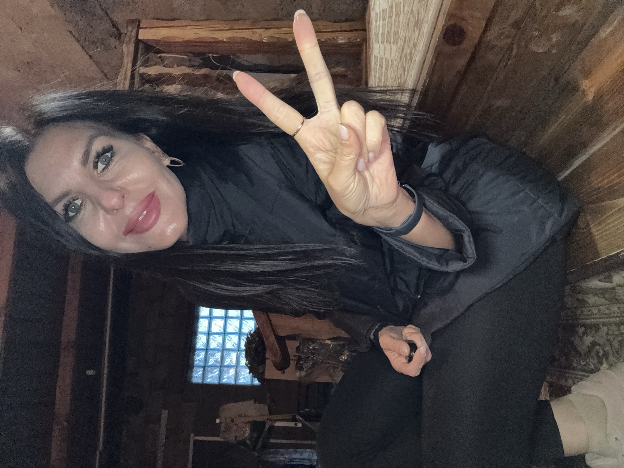 A woman with long, dark hair and light-colored eyes poses indoors, giving a peace sign with her right hand. She is wearing a dark jacket and black pants. Behind her, there is a window made of glass blocks. Wooden walls and ceiling are visible. The location is Dubovaya, Russia.