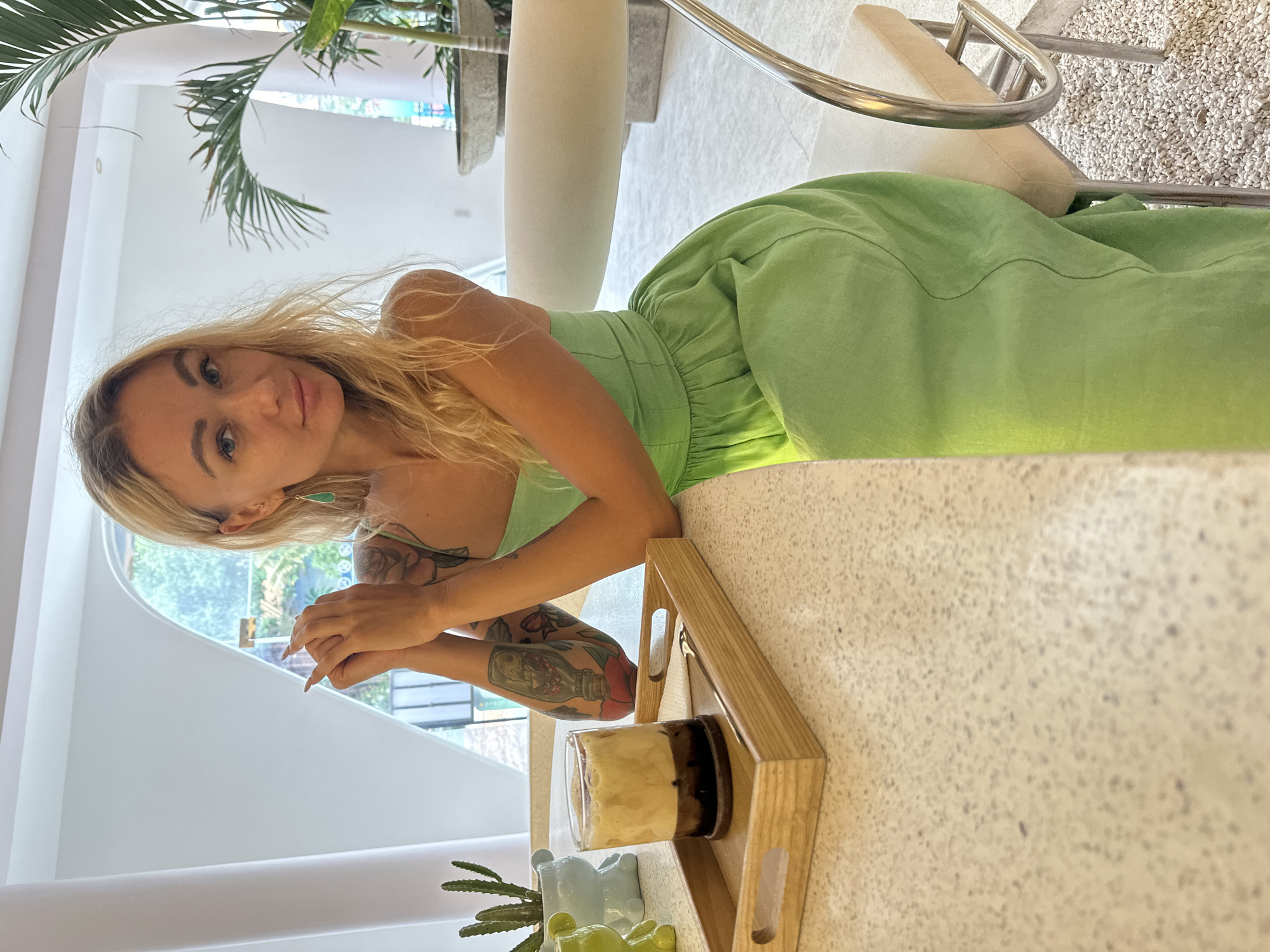 A blonde woman with visible tattoos is seated, looking at the camera in Phu Thuy Ward, Vietnam. She is wearing a light green outfit and rests her arms on a countertop. A wooden tray holds a drink with visible layers and a small cactus in a bear-shaped pot. The environment includes a large window, palm tree, and a modern white chair.