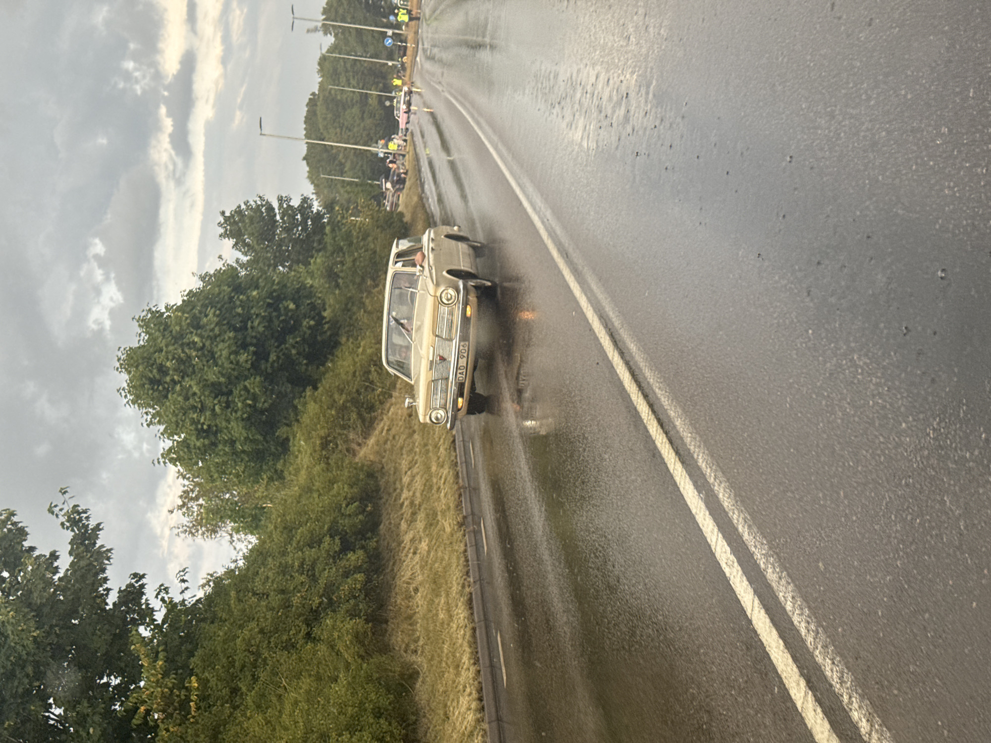 An overcast, rainy day is depicted on a wet, asphalt road with double yellow lines. A classic beige car, possibly a Plymouth Valiant, drives on the right side of the road. The car has the license plate "DAO 906". Several other vehicles and people are visible in the background, indicating a possible event or gathering. Trees and roadside vegetation line the left side of the road, and streetlights are present along the right. The scene is set in Falkenberg, Sweden.