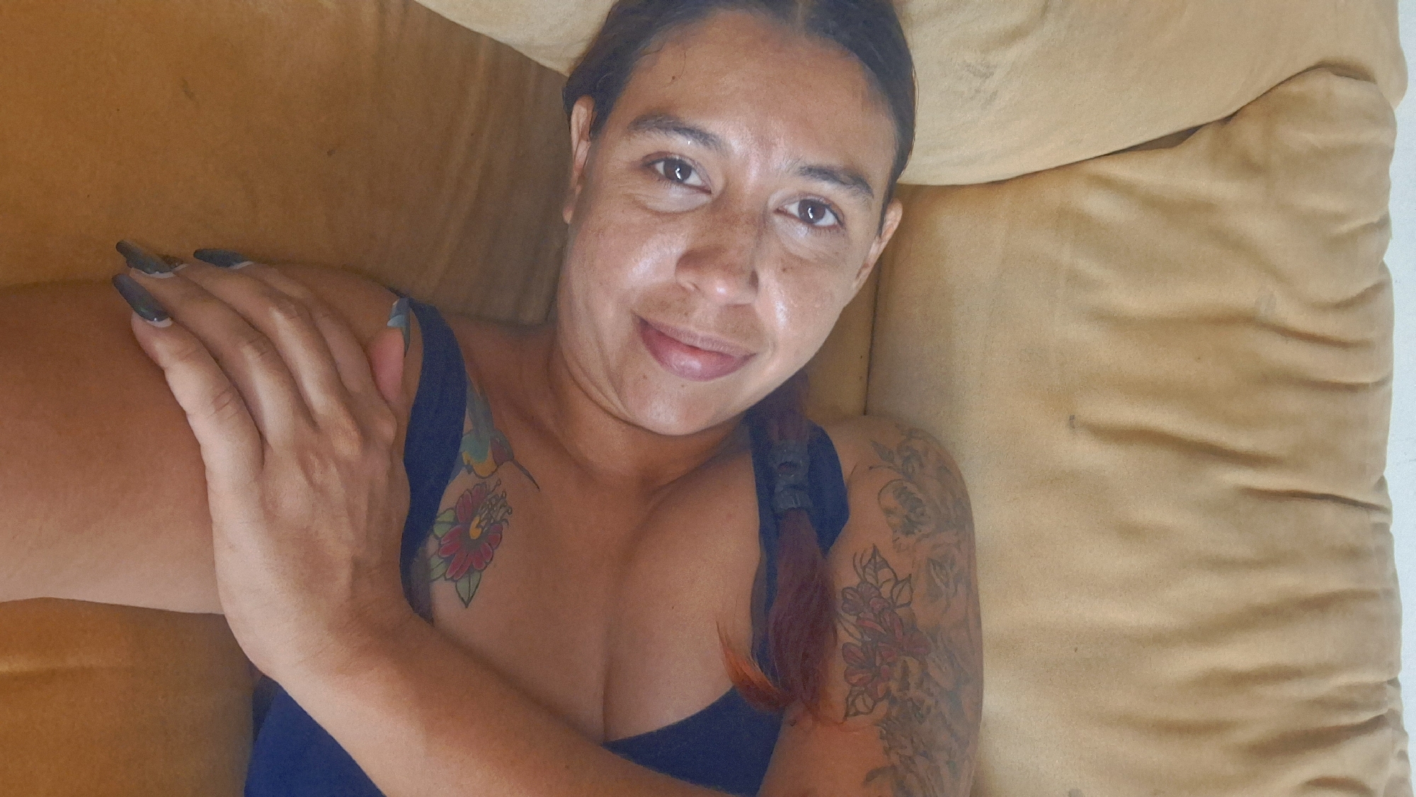 A woman is lying on a beige couch in a room. She is lying on her side with her head resting on her arm and her body is slightly angled towards the camera. She is wearing a blue top and has tattoos on her arms and chest. She has dark hair that is braided and her eyes are looking at the camera. Her lips are slightly parted and she has a small smile on her face. The couch is textured and appears to be made of fabric. The background is slightly out of focus but appears to be a wall. The lighting in the room is soft and natural. The image was taken in Vallecito, Venezuela.