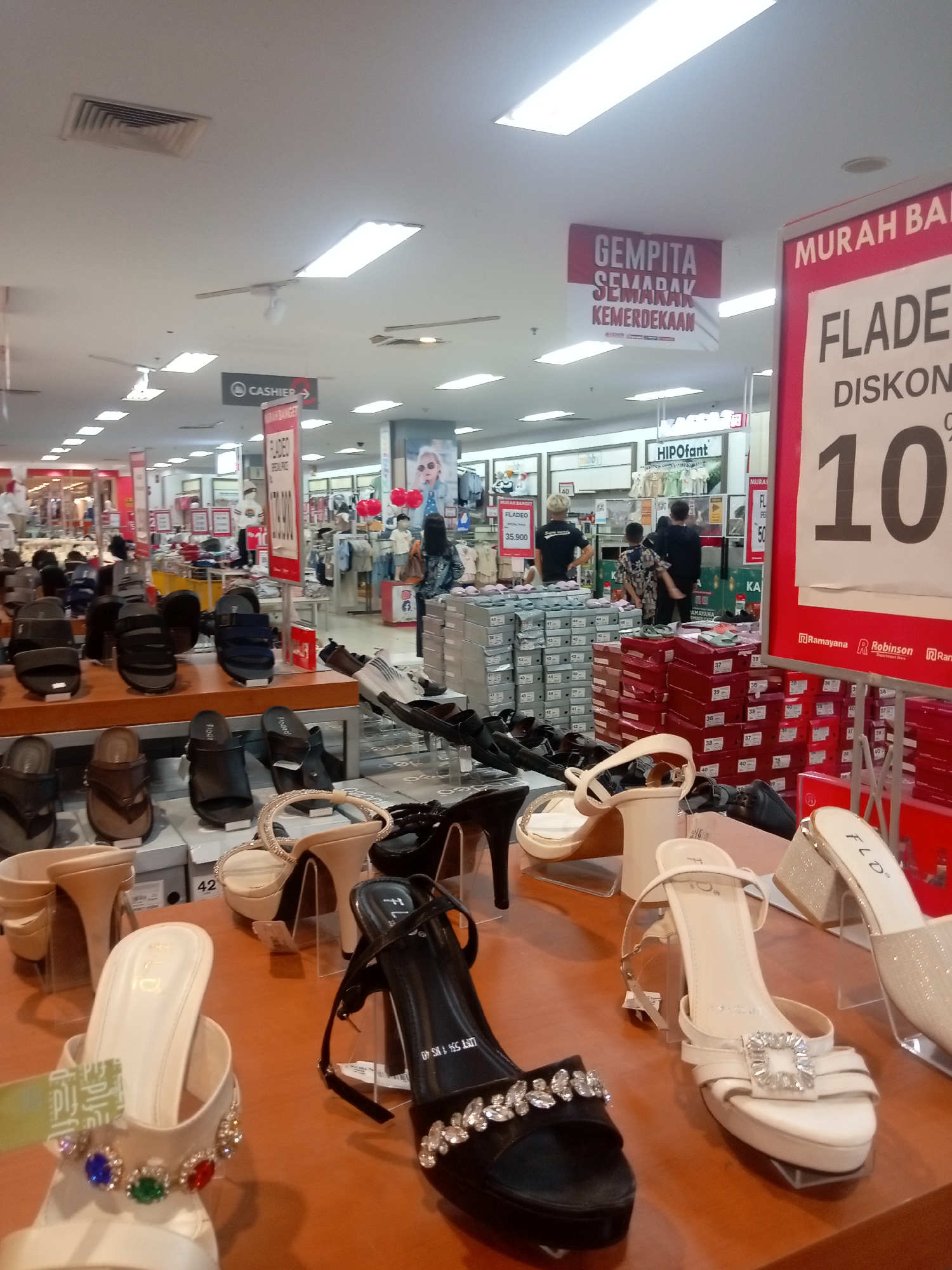 The image shows a retail store, likely a department store, selling footwear. The foreground displays a variety of women's shoes, including high heels adorned with embellishments, platform sandals, and casual slip-on sandals. The shoes are presented on wooden shelves and clear display stands. In the mid-ground, more shoes are visible on shelves, along with stacks of shoe boxes. A sign above reads "CASHIER" with an arrow pointing to the right. Further in the background, clothing racks and mannequins suggest a broader retail selection. Two large banners are visible hanging from the ceiling. One, in red and white, reads "GEMPITA SEMARAK KEMERDEKAAN", which translates to "Festive Independence Day". Another banner, predominantly red, features the text "MURAH BARU FLADE DISKON 10%" indicating a sale with a 10% discount. Brands like "FLP" and "Ramayana Robinson Department Store" are visible on products and signage. The environment appears to be an indoor shopping mall or a large retail space, with bright fluorescent lighting and a tiled floor. The presence of a cashier sign and varied merchandise strongly suggests this is a commercial establishment in Batam, Indonesia, likely within a shopping center.