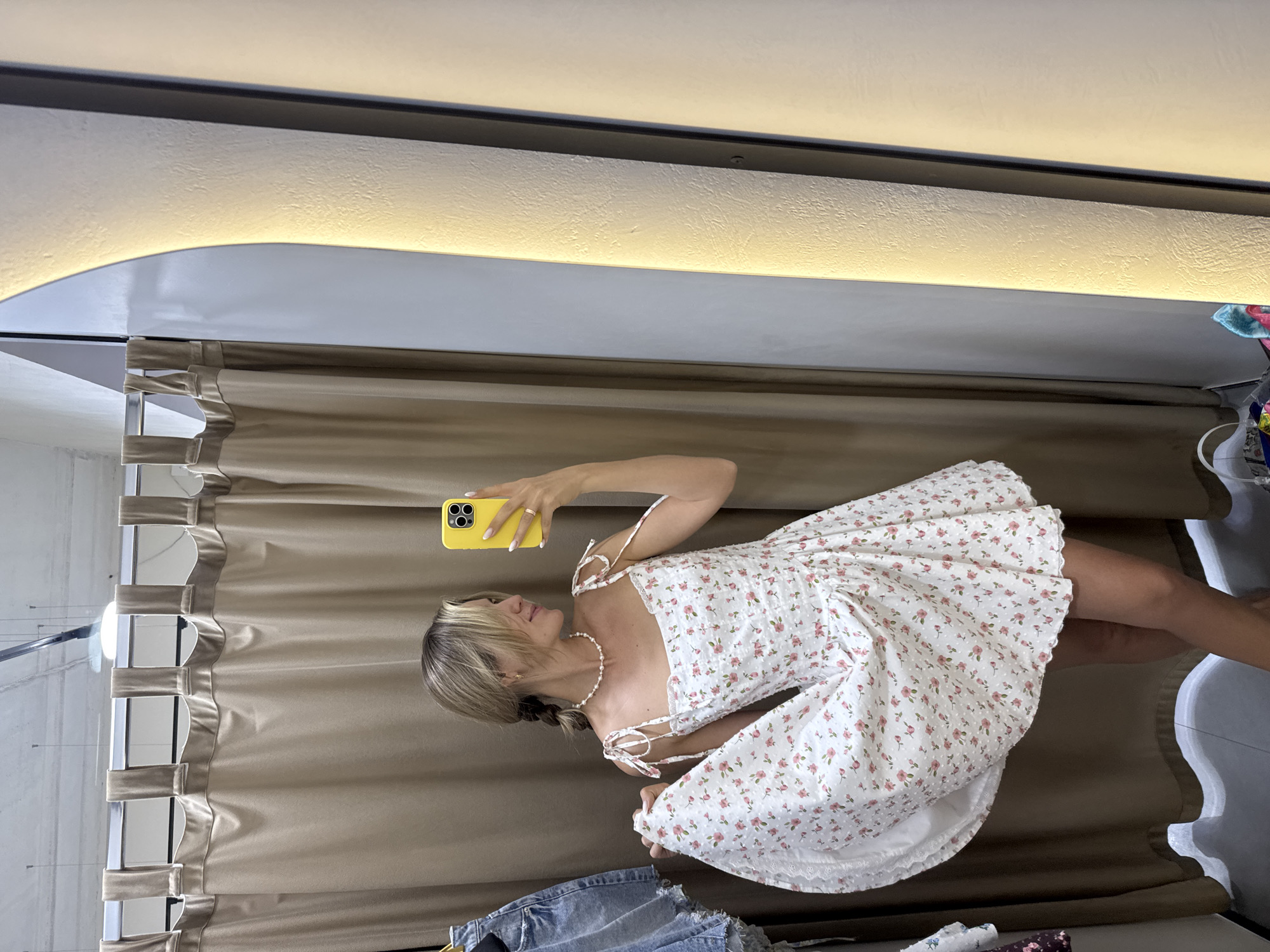 A young woman with blonde hair in a braid is standing in a fitting room and taking a selfie in a mirror. She is wearing a white dress with a floral print of pink flowers and small white dots. The dress has a square neckline and tie straps. She is holding up the hem of the dress with her left hand, showing off its full skirt. Around her neck, she is wearing a pearl necklace. Her phone, which she is holding to take the picture, is yellow. The fitting room has beige curtains that are partially drawn. There is a light fixture above the curtains. To the left of the woman, a pair of denim shorts is hanging on a rack. To the right of the woman, a tall, vertical mirror with a warm light strip is visible. The floor is a light grey.