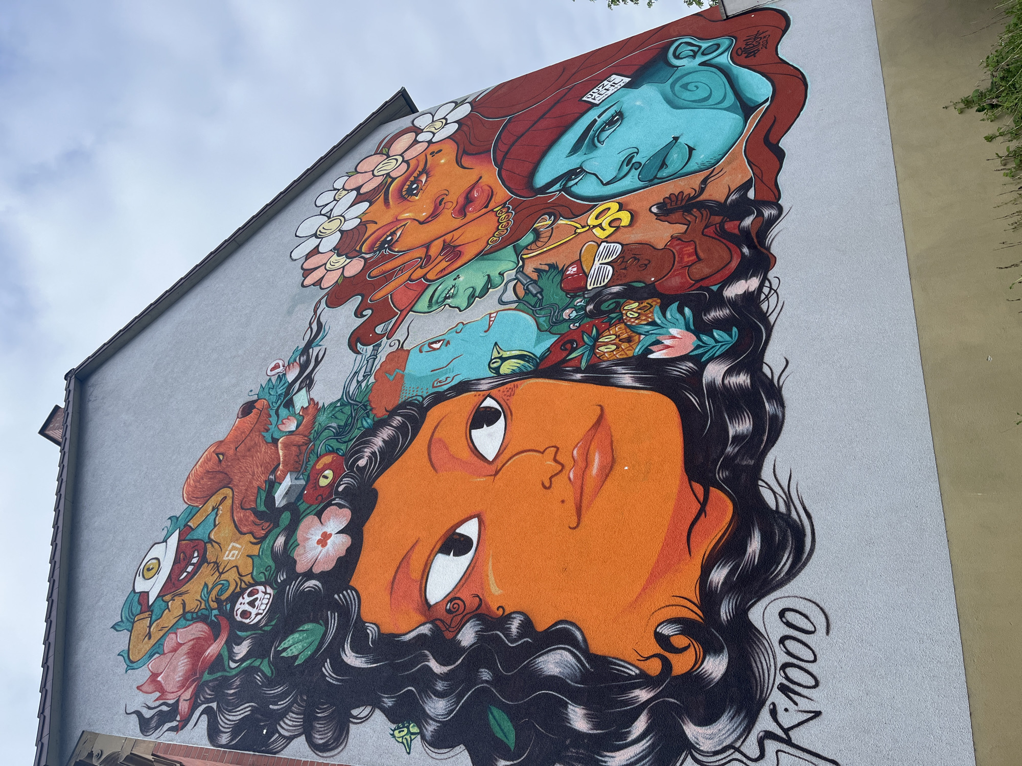 A large, vibrant mural adorns the side of a building, depicting a series of stylized faces and motifs. The mural is dominated by a large, orange-skinned face with distinct features and flowing black hair. Above this face are several other faces, each with a unique color palette and expression. These faces are interspersed with various objects, including flowers, animals, and other abstract shapes. The building is located in the city of Kehl, Germany. The building's exterior is painted a light gray color. The mural itself is brightly colored and features a variety of different artistic styles.