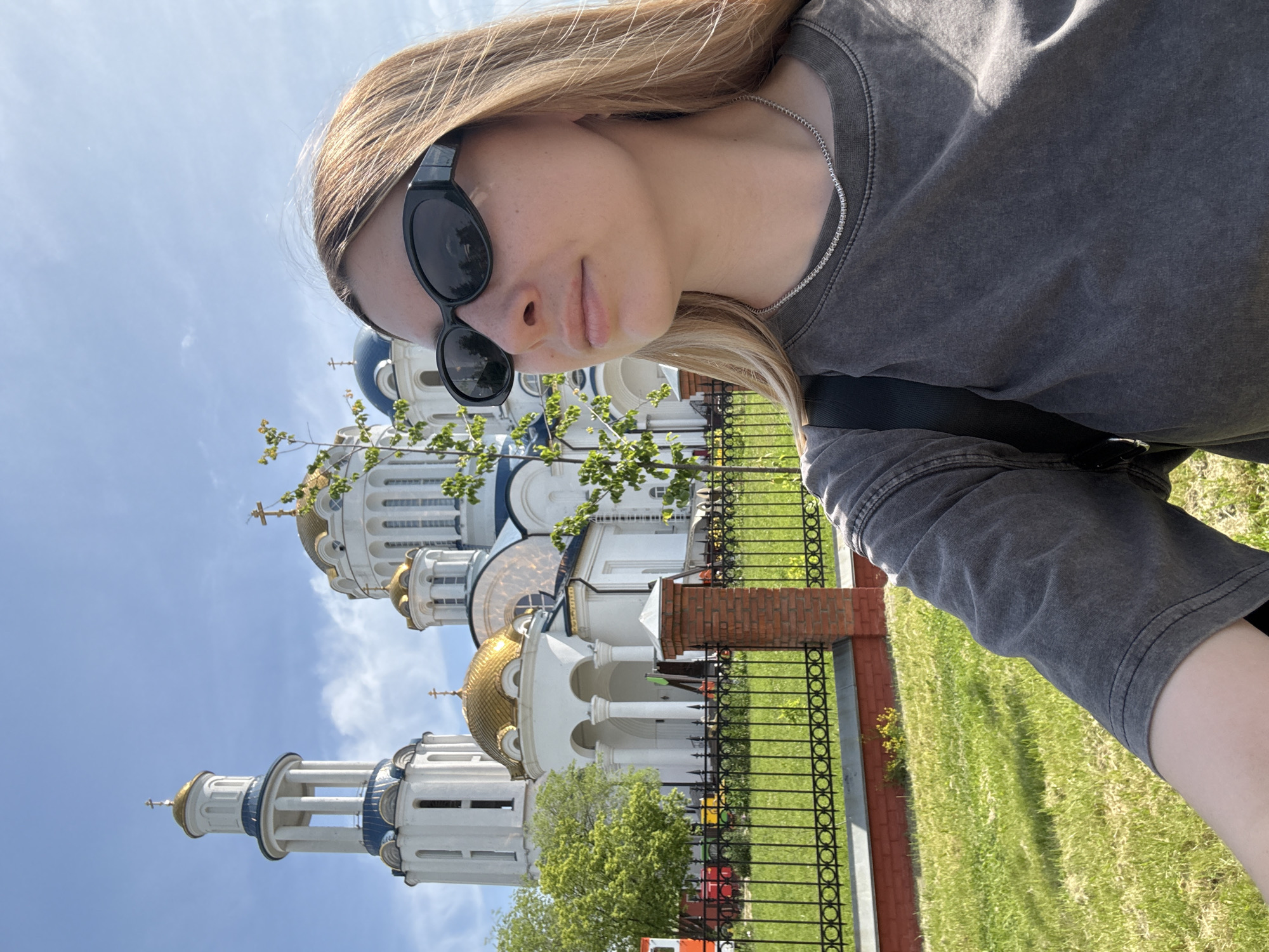 In an outdoor, daytime, selfie-style photograph, a young Caucasian woman with blonde hair and sunglasses is positioned in the foreground, looking to her right. She is wearing a dark gray t-shirt and a silver chain necklace. Behind her, a large, white, domed church with gold accents and blue trim is visible under a clear blue sky with scattered clouds. A black metal fence runs horizontally across the middle ground, separating the woman from the church. Lush green grass and trees are present in the foreground and midground. The location is identified as Moscow, Russia.