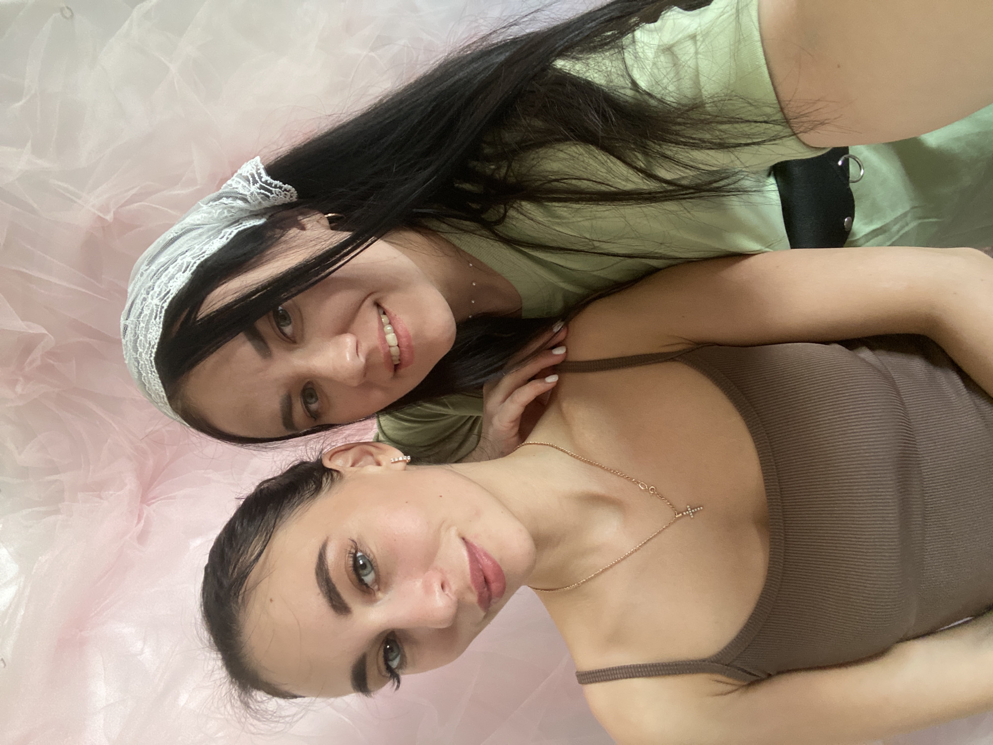 Two women are lying on a pink tulle fabric. One woman is wearing a light green t-shirt and a white lace headband. The other woman is wearing a brown tank top and a gold necklace with a cross pendant. The background is pink tulle. The location is unknown.