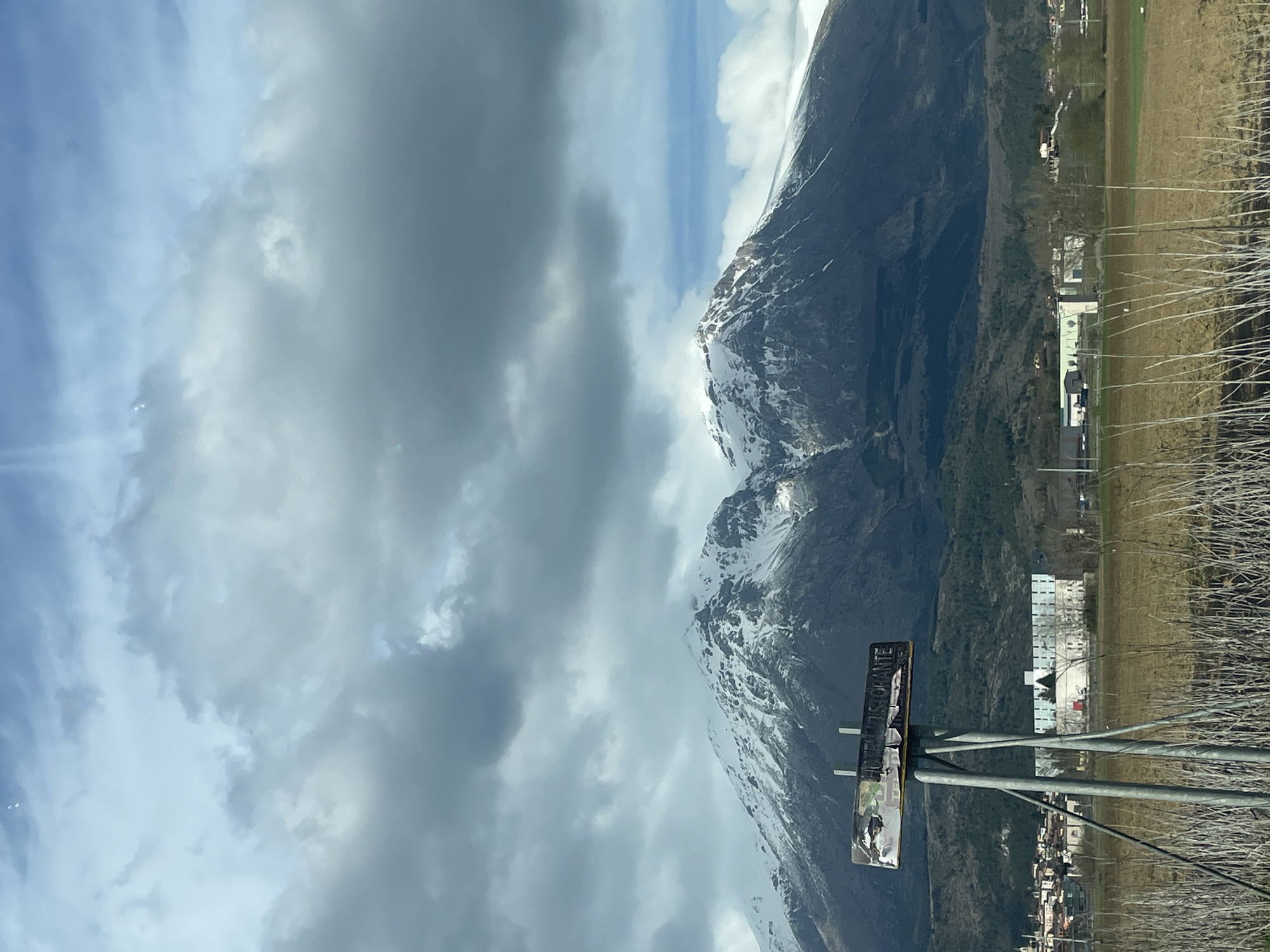 The image shows a view of a mountain, likely taken in Scurcola Marsicana, Italy, based on the presence of buildings consistent with the region's architecture and the general landscape. The mountain is partially covered in snow. To the right of the mountain, there's a view of the land and architecture of the local town of Scurcola Marsicana, with buildings scattered across the landscape. There's a sign on a pole that reads, "Ristorante" is situated in the foreground. The sky is filled with cloudy weather. Bare branches, likely trees, also appear in the foreground, obscuring the buildings behind them.