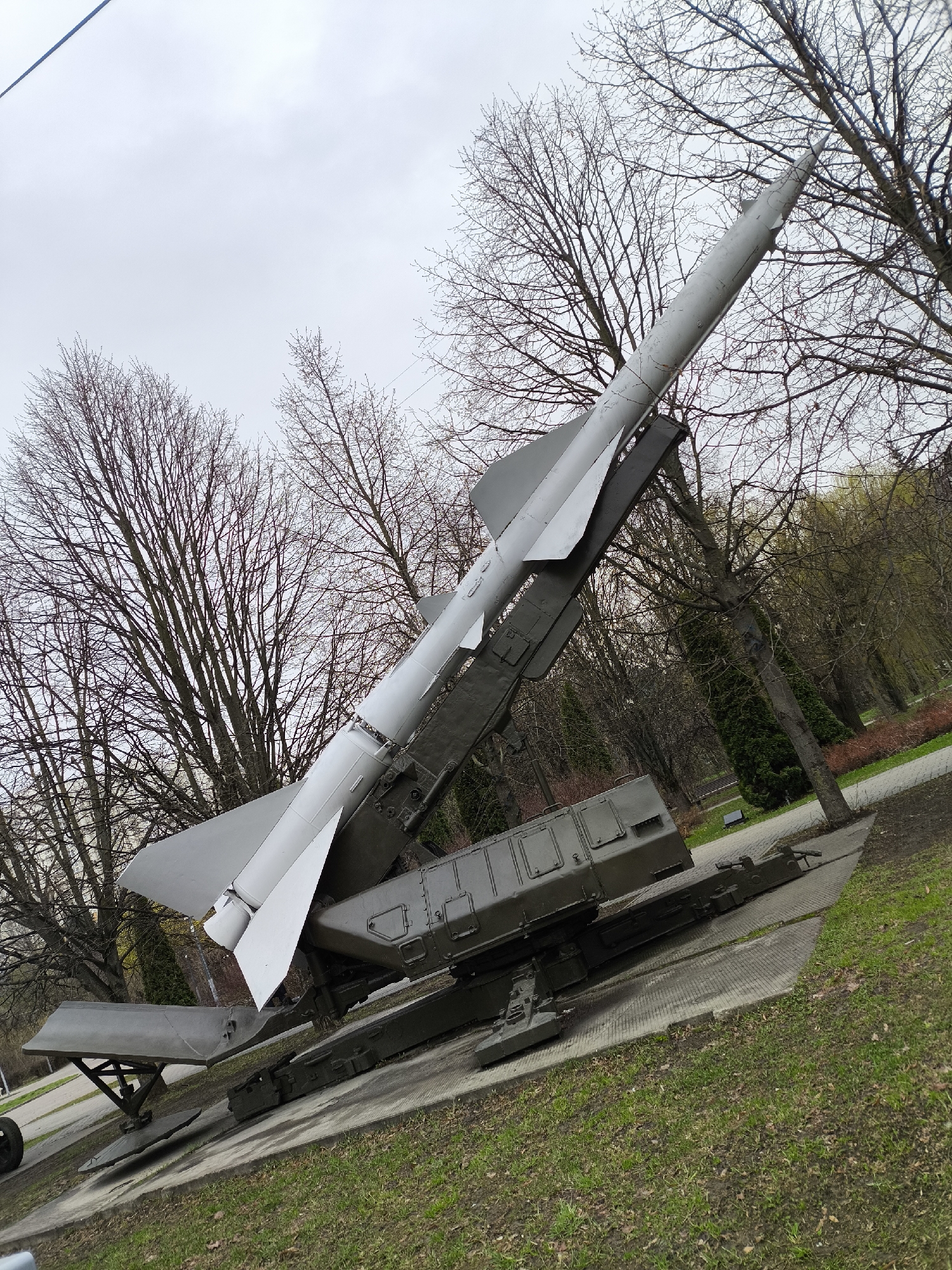 The image displays a large, light grey missile mounted on a dark green mobile launch platform, situated outdoors in what appears to be a park or open-air museum in Khimki, Russia. The missile features a long, conical nose, a cylindrical body, and prominent delta-shaped fins at its base. It is angled upwards, as if in a launch-ready position.
The robust launch platform, which supports the missile, includes various structural components and what appear to be equipment housings or control units at its base. The entire display rests on a grey concrete or paved area, surrounded by patches of green grass and bare soil.
The background is dominated by numerous deciduous trees with intricate bare branches, indicating late winter or early spring. Further back, paved pathways weave through the park, alongside some evergreen vegetation and other trees that are beginning to show hints of green. The sky is overcast and a bright, uniform grey, providing diffused and flat lighting to the scene.
No people are visible in the image, and there is no discernible text on the missile or its launcher. The scene conveys a quiet, solemn atmosphere, characteristic of a static historical military exhibit. The missile itself, given its design and location, likely represents a Soviet-era surface-to-air missile such as an S-75 Dvina (SA-2 Guideline).
