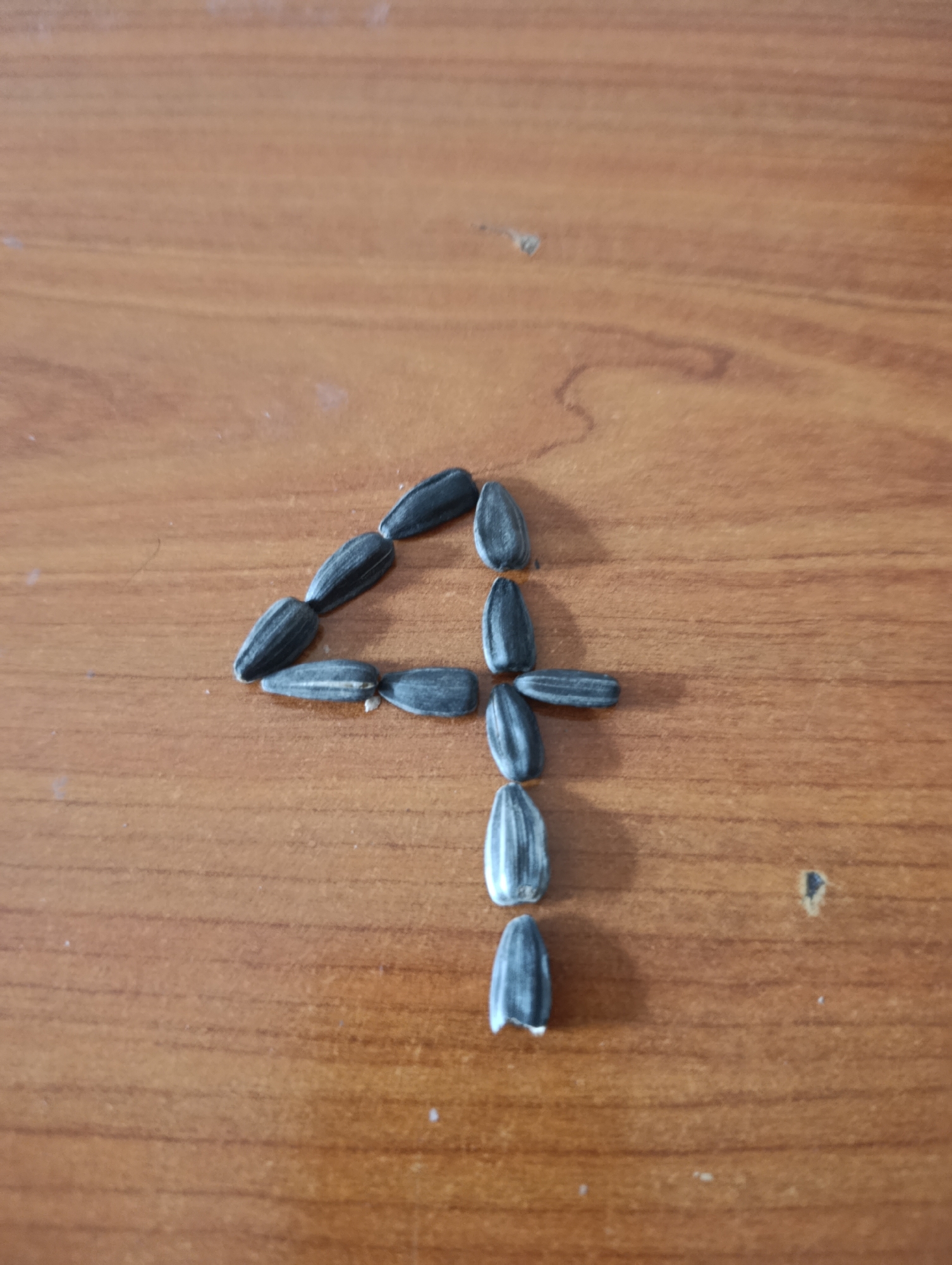 The image depicts the number "4" constructed out of sunflower seeds on a wooden surface. The wood appears to be a table or desk with a clear grain pattern. The seeds are predominantly black with some showing white stripes. The arrangement forms a recognizable numeral four. The environment appears to be indoors, possibly a home or office setting. No people are visible. The location is not discernible from the image alone.